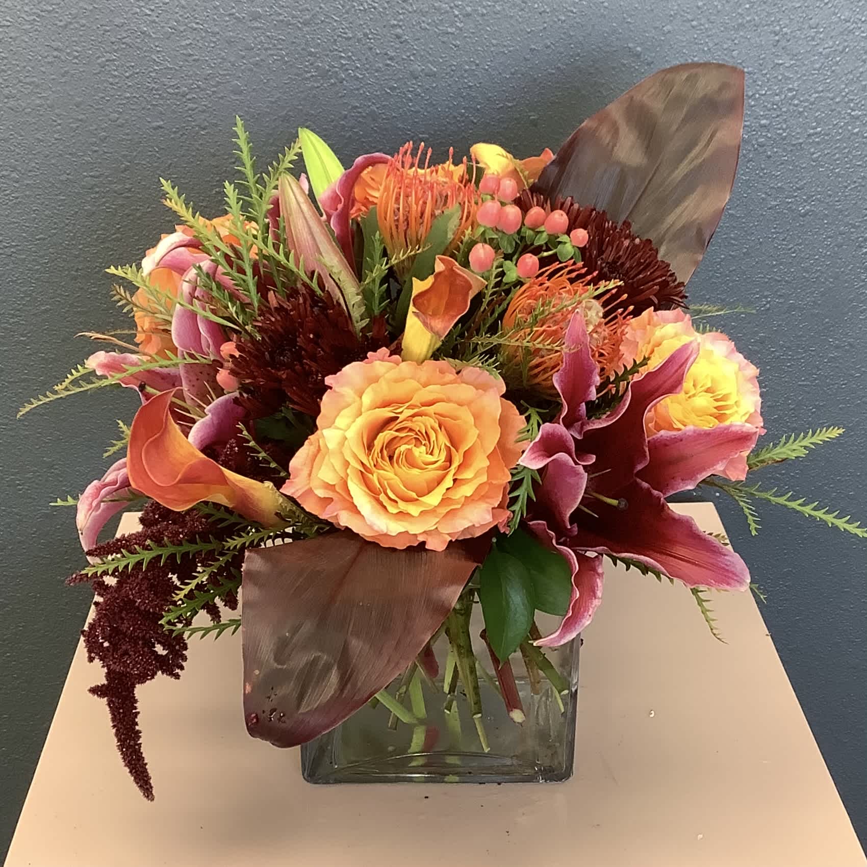 Autumn Love - When you want to send the very best to that special loved one. Featuring stunning premium blooms such as, protea, roses, lilies and much, much more. All designed in a cube vase, this breathtaking arrangement is perfect for your holiday table.   This arrangement can be customized for any pallet or occasion. Flowers and container may vary, please call if this is a concern.