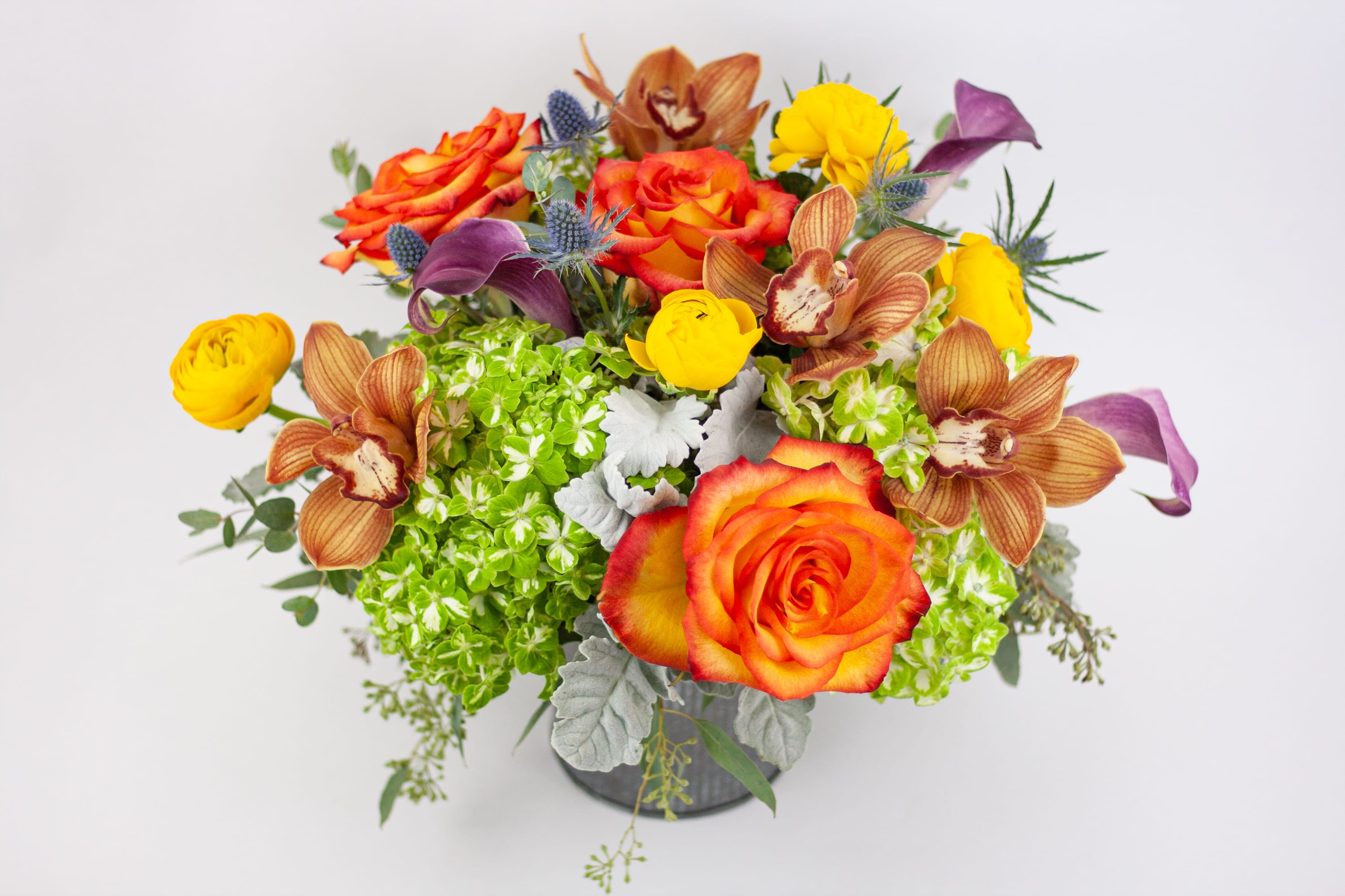 Wild Sunrise  - BRIGHT VASE OF FLOWERS WITH CYMBIDIUM ORCHIDS AND ROSES   Bright, creative and eclectic. A gorgeous vase of vibrant mixed blooms with interesting shapes, textures and colors. Tropical cymbidium orchid blooms star in this artful design, nestled with frilly yellow ranunculus and bright bi-color ‘High Magic’ yellow roses with red tips. Tropical burgundy-purple calla lilies, pops of green hydrangea and spiky silver-blue thistle complete the look.  These flowers are perfect for folk with creative tastes who like to stand out in a crowd.  Send this vase of flowers to wish someone a special happy birthday or a get well soon. Deliver to a client or colleague in recognition of an achievement or as a thank you.  Why not add a greeting card or quality handmade candle to match the occasion or personality?  Wild Sunrise features: ·        Orange cymbidium orchids ·        Bi-color ‘High Magic’ yellow-red roses ·        Bright yellow ranunculus ·        Green hydrangea ·        Silver Dusty Miller foliage ·        Burgundy-purple calla lilies ·        Silver-blue thistle ·        Seeded eucalyptus ·        Metal ribbed vase   SIZE: The STANDARD flower arrangement is approximately 10” height and 15” width.   ALLERGY ADVICE: This design has low pollen and a low fragrance. Calla lilies are known to be mildly poisonous if ingested by cats or dogs.   FAQ Q: What time will my flowers arrive and what if no-one is there to accept them? A: Excellent question! We’re unable to provide or guarantee specific delivery times. All our flowers are delivered anytime between 9-5pm, with exception to Sympathy designs for a funeral service. For full details around our delivery process and timings, please see our Terms and Conditions. Q: What days do you deliver flowers? A: All our flowers are available for San Francisco and Daly City delivery seven days a week, between 9am-5pm. Q: May I order for the same day delivery? A: Yes, same day deliveries are available if you place an order before 1pm. Q: May I request to change the design a bit? A: Yes, please make a note in the SPECIAL INSTRUCTION box when you place your online order. We will do our best to follow them. Q: Will my flowers look exactly as what is pictured? A: Depending on the season, colors or shades may vary. Sometimes we need to substitute flowers due to seasonal availability, but they’re always substituted for the same or greater value.   Ready to bring floral beauty into someone’s world with our creative and bright vase of flowers? Order this gift online now or give us a call on (415) 752-2060 if you have a question or special request. 
