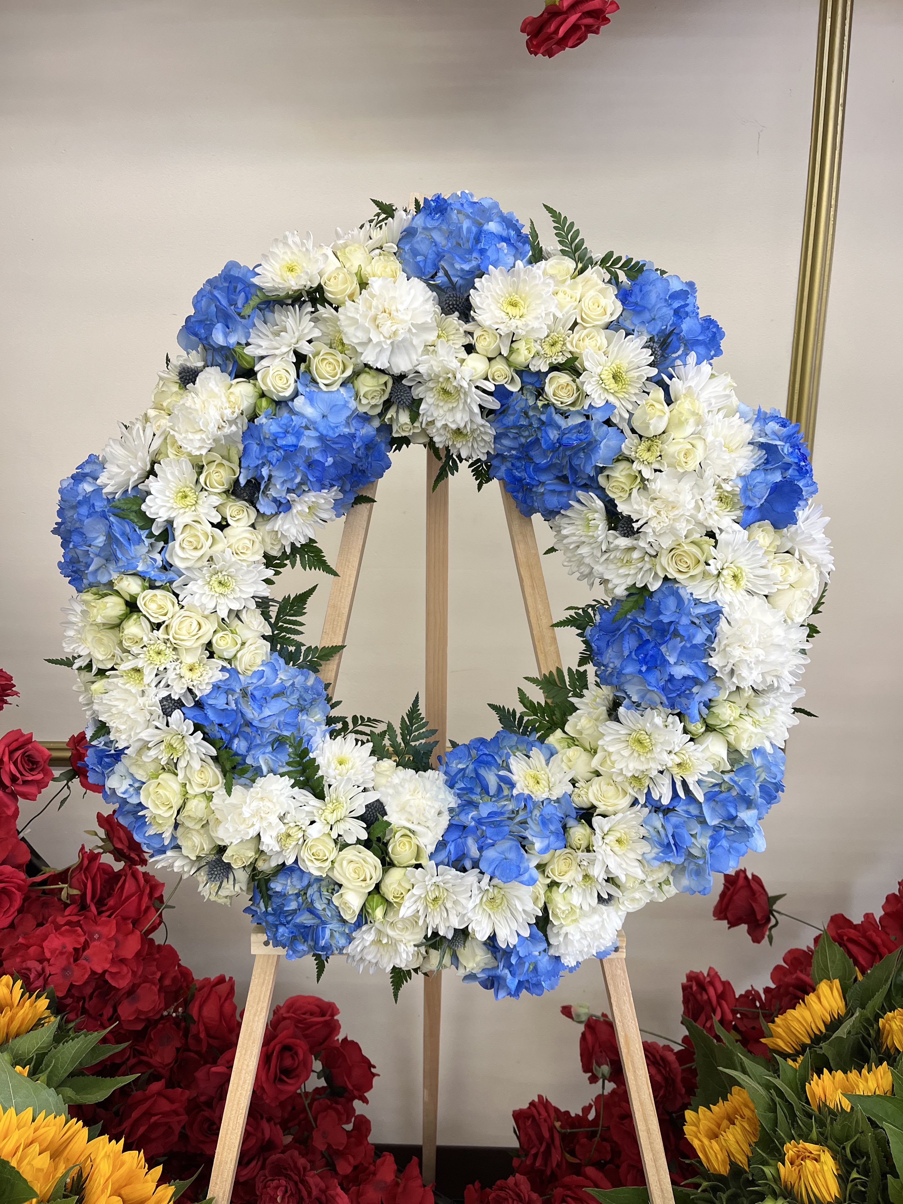 Haven Tribute Wreath - This serene funeral wreath features white roses and cushion blooms paired with royal blue hydrangeas and delicate blue thistle flowers. The calming blend of white and blue creates a graceful and peaceful tribute, symbolizing remembrance and eternal love. **Note: PICTURED IS THE DELUXE SIZE.  Easel is INCLUDED**