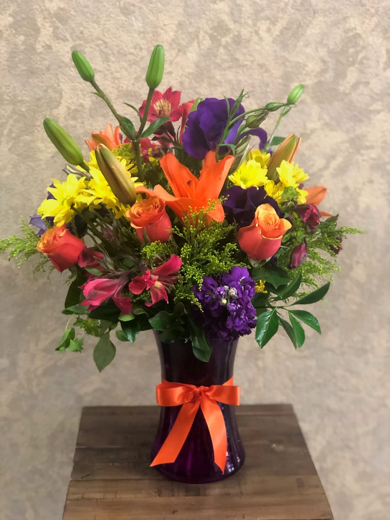 Positano, Italy - Type of Flowers: Purple Stock, Coral Roses, Orange Lilies, Purple Lisianthus, Pink Alstroemeria, and Yellow Daisies in a purple vase with orange ribbon. Availability: All year round Substitute Available: Yes Design View: All-sided Photo shown: Regular