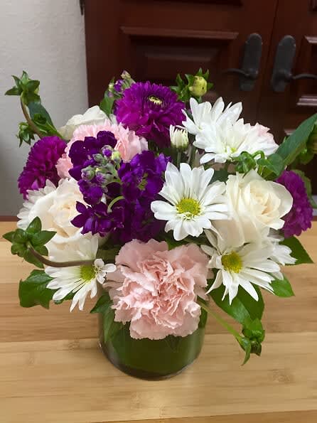 Sweet Daisies - Type of Flowers: White Roses, White Daisies, Purple Stock, Pink Carnations, Cushion Mums, and Greens in a small clear vase with leaf wrap inside. Availability: All year round  Substitute Availability: Yes