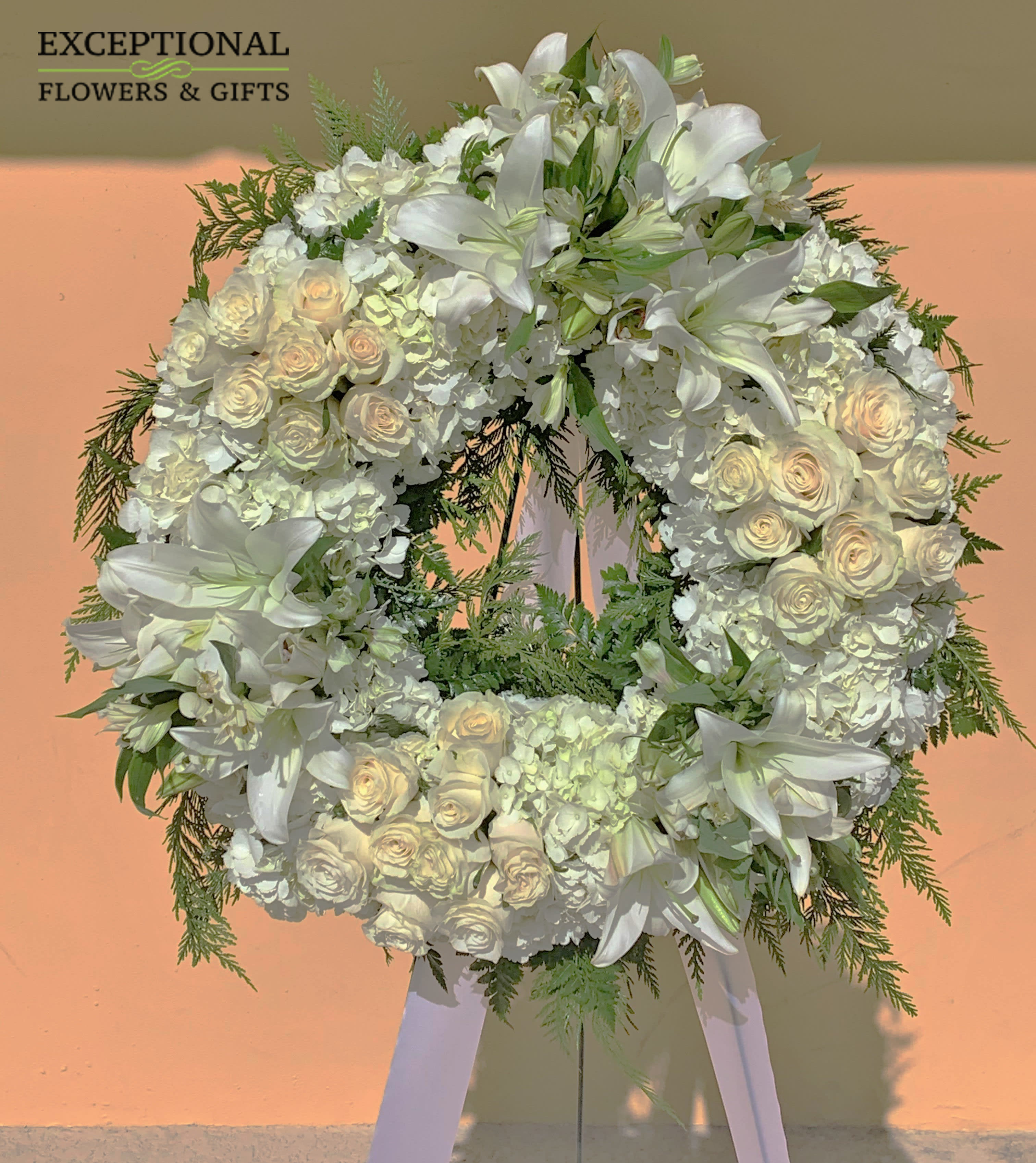 Wreath of Remembrance (BR) - The traditional funeral wreath, beautifully representing the eternal circle of life, is updated for a new era and is beautifully refreshed without losing any of its poignant meaning. This impressive wreath is constructed of white roses, carnations and Asiatic lilies complemented by lush greens and ribbon and trim. It makes a superb, time-honored addition when displayed on a wooden easel for a wake or funeral service, graveside at a burial, to decorate a headstone or to set adrift for burials at sea.   