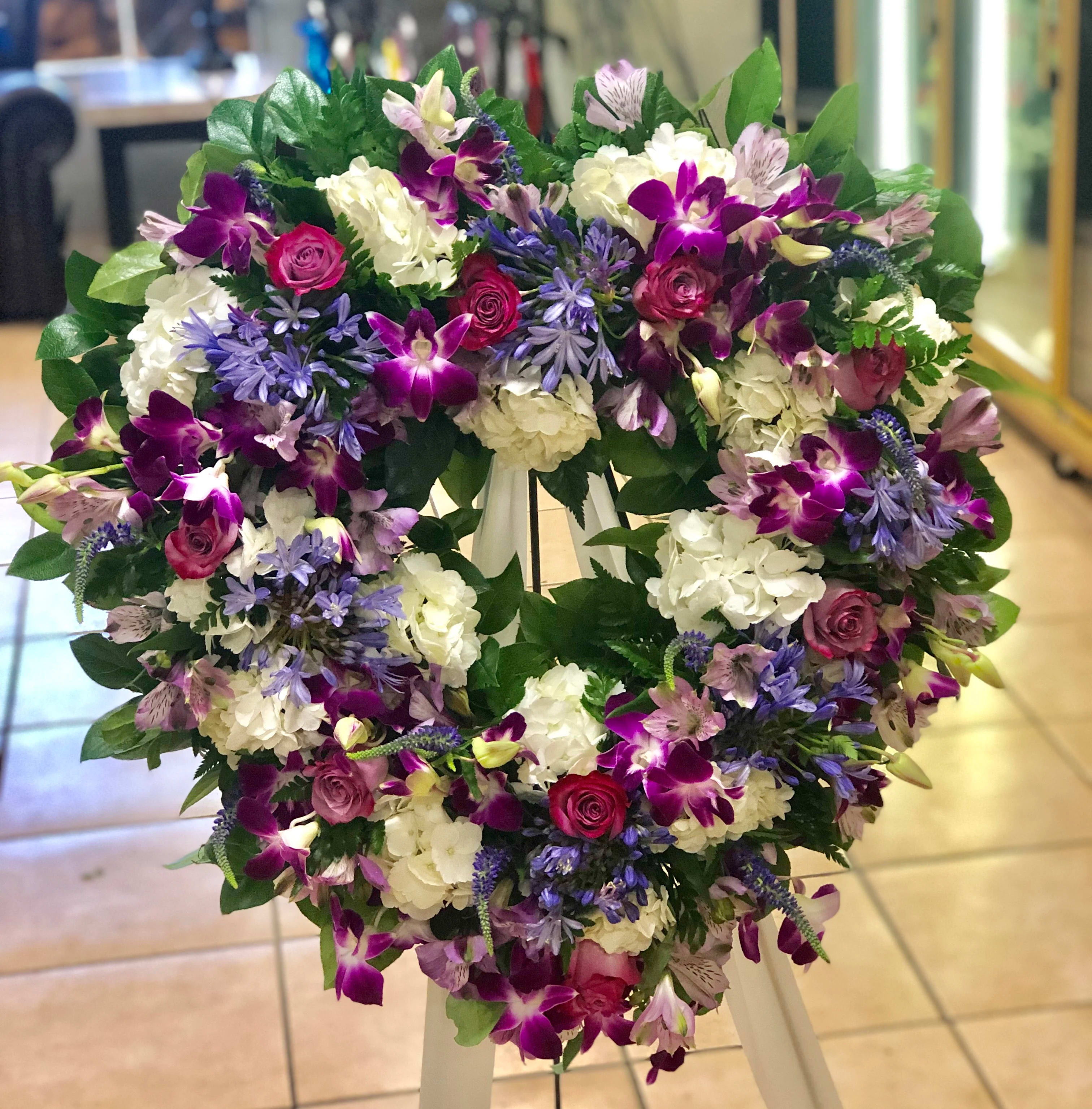 Heavy Heart (BR) - Presenting our exquisite open heart standing spray, a heartfelt tribute overflowing with beauty and grace. This stunning arrangement combines a selection of enchanting flowers, including purple roses, purple dendrobium orchids, white hydrangea, agapanthus, purple veronica, and lush greenery.
