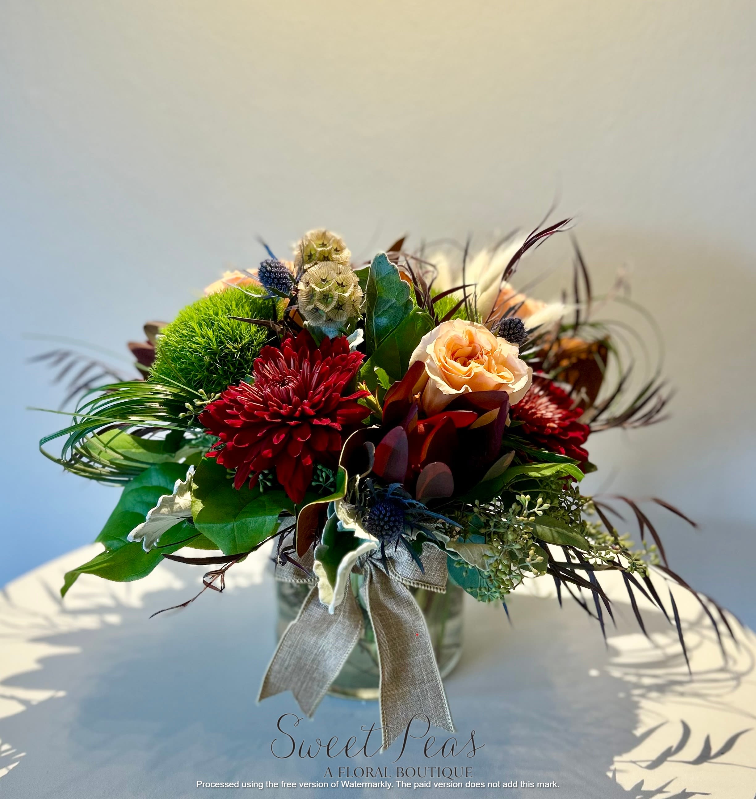 Amber &amp; Merlot - A lovely mix of textures: warm peach roses, rich burgundy mums, earthy pods, spiky thistle, soft grasses, and a natural bow grounding it all. The palette feels autumnal yet elegant, with a balance of rustic charm and sophistication.