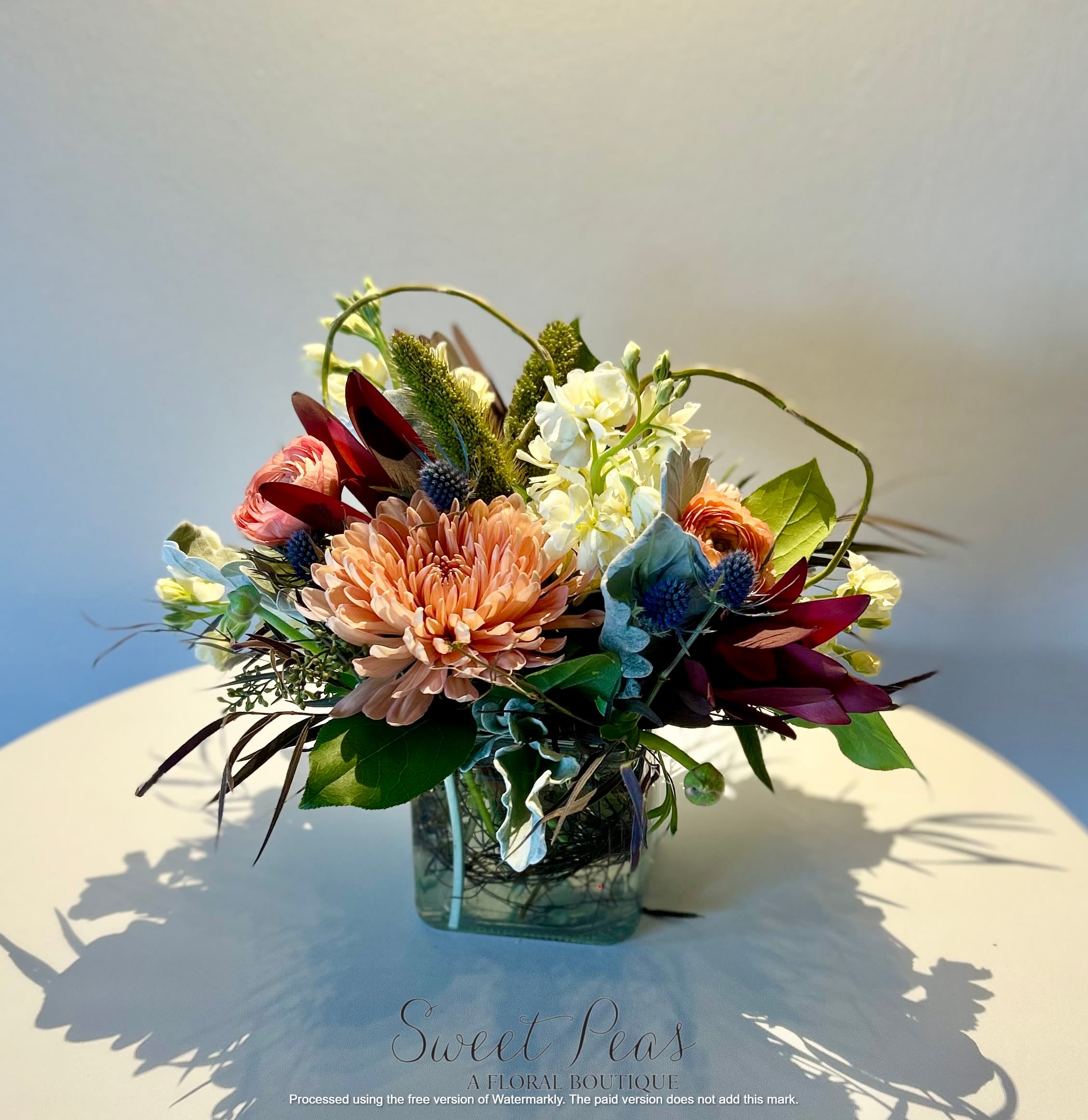 Sunset Whispers - Soft and airy—peach mums, creamy stock, soft ranunculus, blue thistle, and muted foliage. It has a light, romantic, and almost painterly quality with a touch of whimsy from the curved grasses.