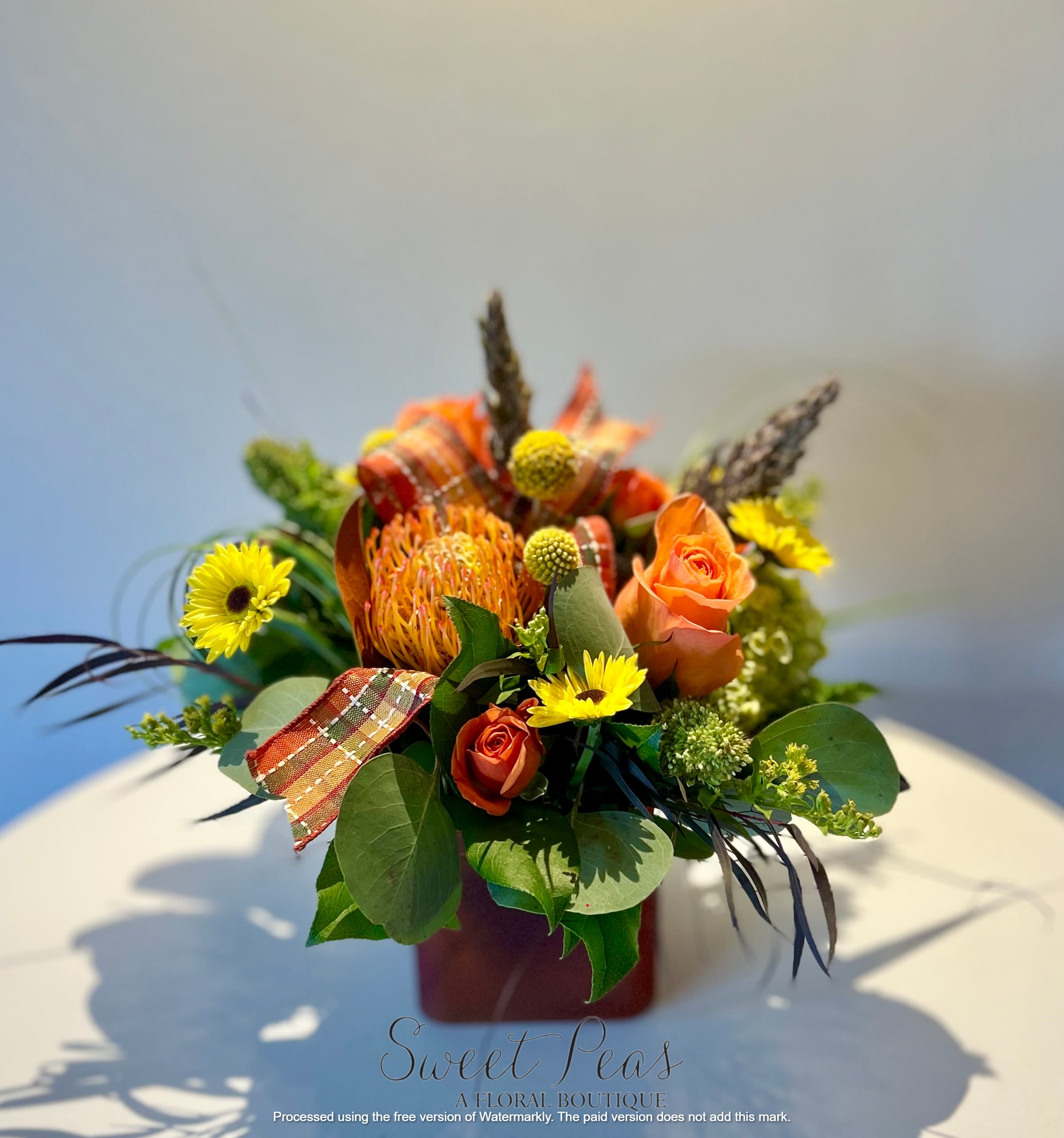 Sunny Harvest - Radiates cheerful warmth—sunny yellows, vibrant oranges, bold protea, playful craspedia, and plaid ribbon for a rustic harvest feel. It’s bright, festive, and full of personality.