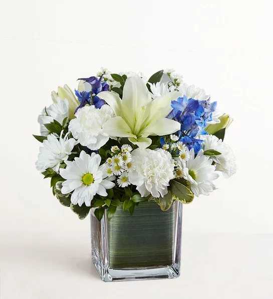 Healing Tears Blue &amp; White - Soothe their tears as they mourn the loss of a loved one with the serenity of blue and white. Our sympathy arrangement of fresh blue delphinium, white roses and lilies, expertly gathered together in a clear glass cube lined with Ti leaf ribbon, makes for an exquisite gesture of comfort and healing.