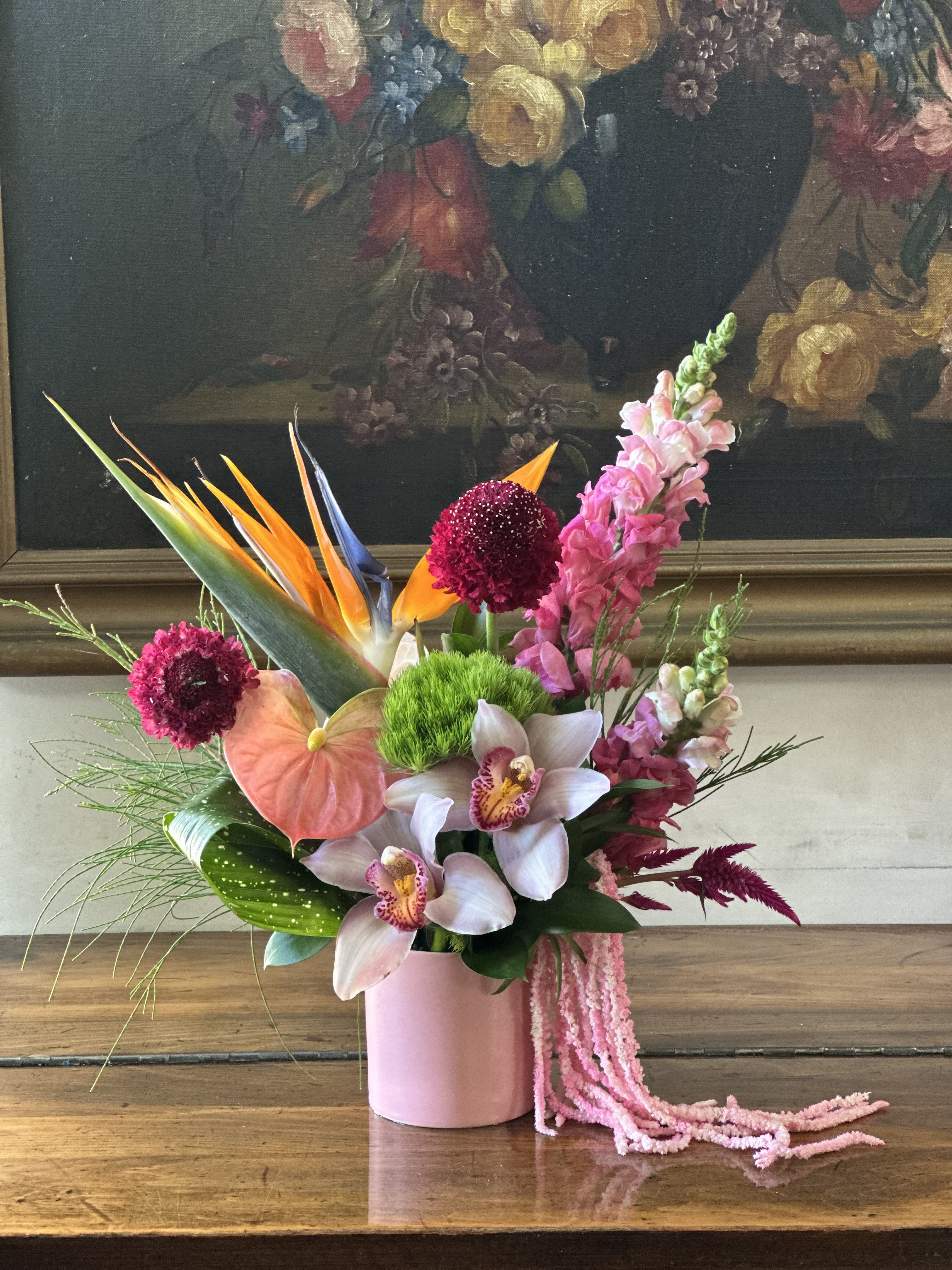 Tropical Coral Reef Reverie  - This exquisite tropical bouquet captures the essence of paradise with its vibrant mix of exotic blooms in a sophisticated pink ceramic vessel. The dramatic bird of paradise takes center stage with its striking orange and blue petals, while delicate white orchids with golden centers add an elegant touch. Rich burgundy dahlias provide depth and contrast, complemented by coral anthurium and soft pink snapdragons that create beautiful vertical lines. Textural elements include bright green chrysanthemum poms and burgundy celosia, while tropical foliage and grasses add natural movement. The arrangement is finished with cascading pink chenille details that echo the vessel’s rosy hue, creating a cohesive and luxurious presentation against the classical backdrop. This tropical bouquet perfectly balances exotic boldness with refined elegance, making it ideal for sophisticated spaces that want a touch of paradise.​​​​​​​​​​​​​​​​