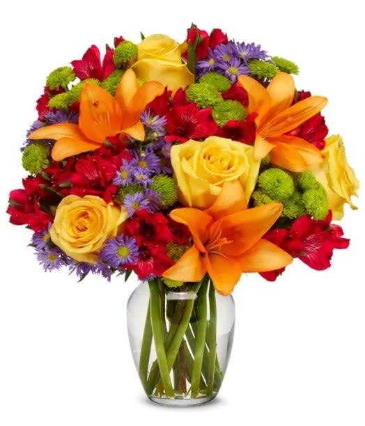 Festive Arrangement  - Arrangement includes orange lilies, yellow roses, red alstroemeria, purple montecasino, green pompon button, and greenery in a clear vase. Picture is  a DELUXE.