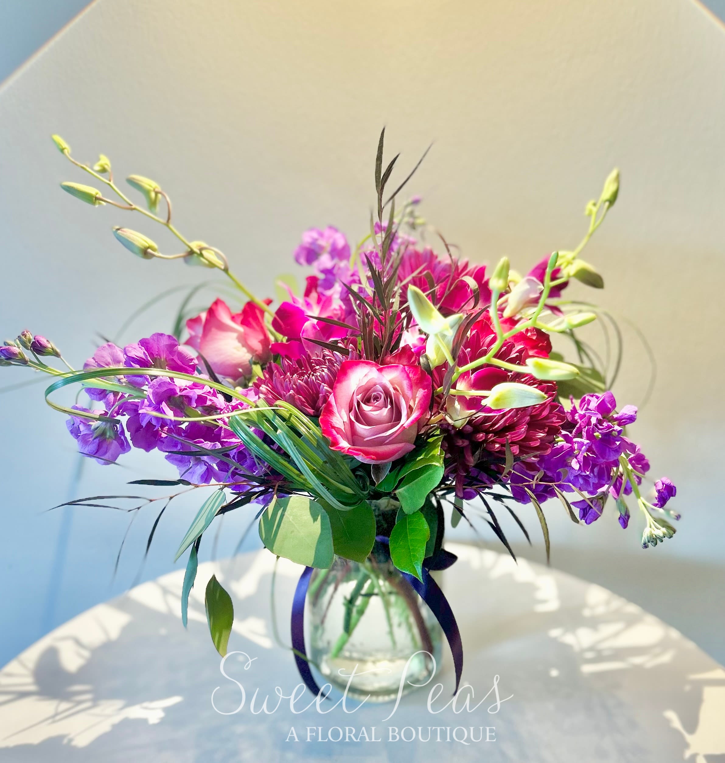 Midnight Orchid - A lush, dramatic mix of fuchsia orchids, magenta mums, violet stock, and bi-colored roses, gathered in a clear glass vase and finished with a sleek ribbon. Midnight Orchid is both romantic and striking, blending deep jewel tones with graceful textures for a look that’s as captivating as it is elegant. Perfect for making a bold statement of love, celebrating a special milestone, or simply bringing vibrant beauty into any space.