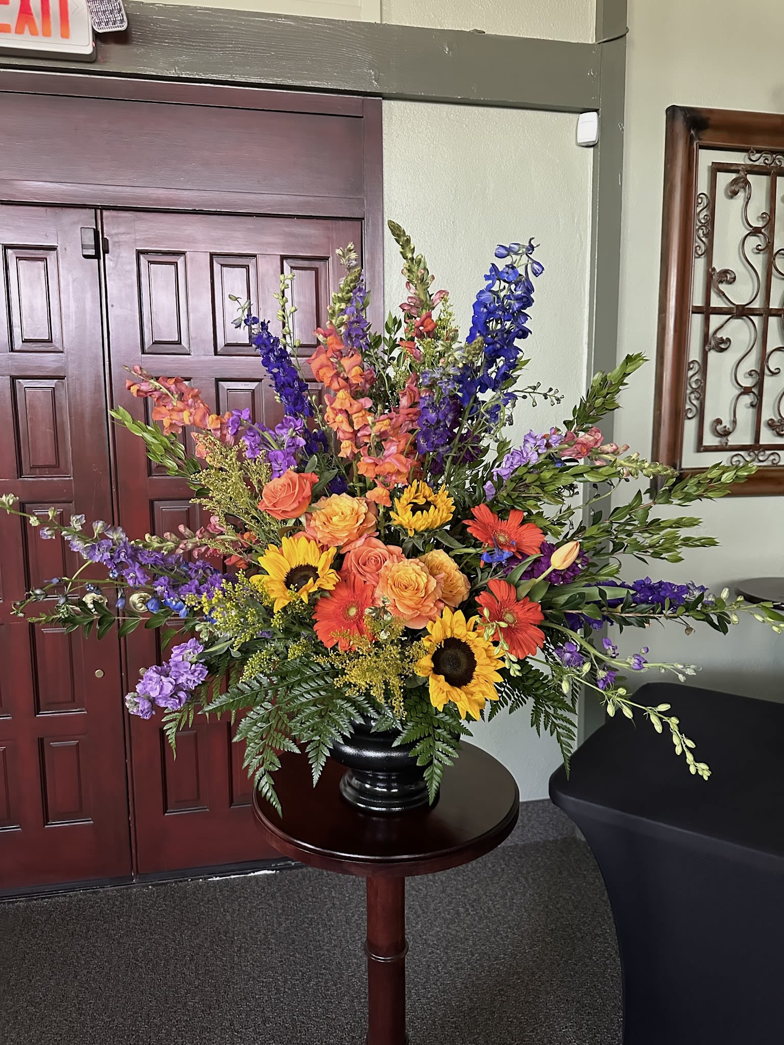 Ceremony Arrangement CV-28 Ceremony Arrangement CV-28 - Celebrate a vibrant life with this striking ceremony arrangement, featuring sunflowers, orange roses, deep purple delphinium, and bursts of coral gerbera daisies. Designed to bring warmth and energy to the funeral service, this funeral floral arrangement honors a joyful spirit with rich color and natural beauty.