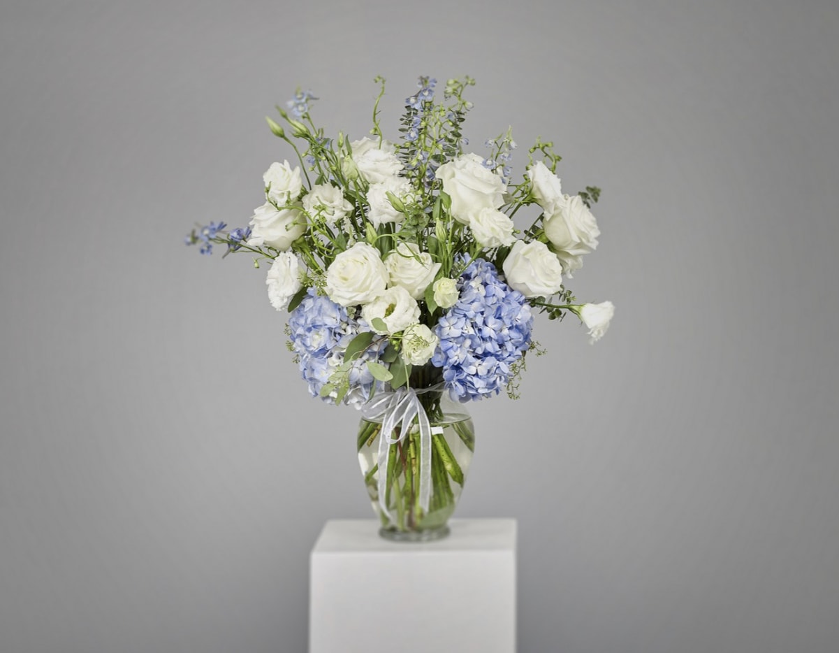 Blue Elegance Blue Elegance - This graceful vase arrangement exudes serenity and sophistication with its soft palette of creamy white roses and blue hydrangeas. Accented with gentle greenery. Each bloom is carefully styled to evoke calm, purity, and tenderness.