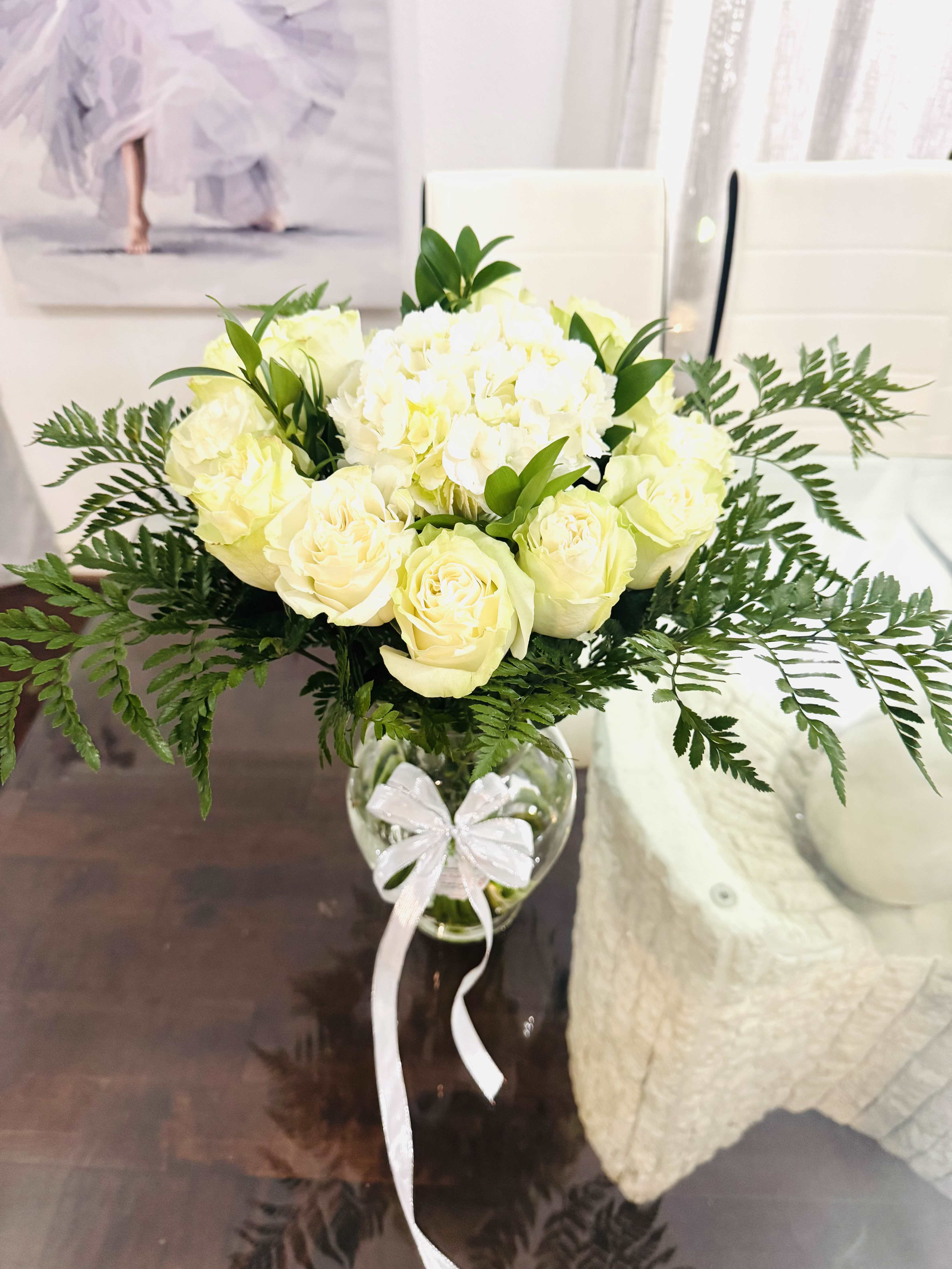Pure Elegance -   Imagine a bouquet where purity and elegance intertwine. At the heart of the arrangement, white roses bloom in velvety spirals — classic, romantic, and timeless. Their creamy petals unfold with a quiet confidence, symbolizing new beginnings and sincere devotion.   Nestled between them, white hydrangeas offer fullness and texture, like soft clouds gathered in gentle clusters. Their delicate florets bring a sense of abundance and grace, adding volume without overwhelming the eye.   Framing the bouquet, leather leaf greenery fans out in rich, deep green tones, its textured leaves adding structure and contrast. The greenery’s bold, glossy presence anchors the lightness of the blooms, making the whites appear even more luminous.   Together, this bouquet is serene yet striking — a natural sculpture of softness and strength, perfect for weddings, sympathy, or any moment that calls for quiet beauty and a touch of the divine.