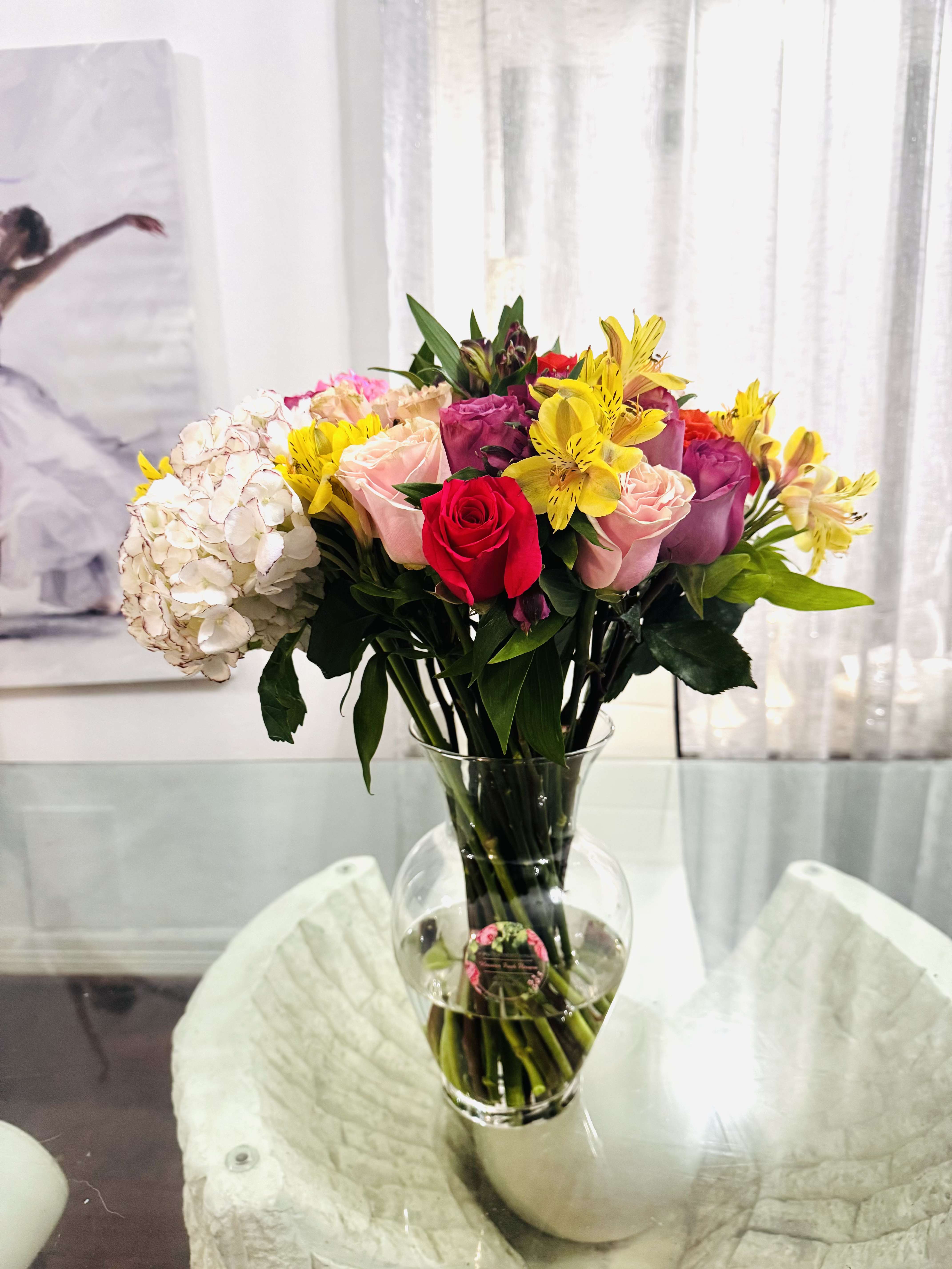 Vibrant Harmony - This bouquet is a radiant celebration of nature's finest artistry- a harmonious blend roses, hydrangeas, and asters, each bloom chosen for its beauty, character, and charm.  At its heart , roses unfold in different layers, bringing classic romance and timeless grace in hues that range from blush to bold. Surrounding them, hydrangeas offer fullness and softness, their cloud-like clusters adding texture and depth, like whispers of early summer skies. Dancing playfully among them, multicolored asters bring cheerful energy and a wildflower spirit- dedicate yet vibrant, like stars scattered across a painter's canvas.  The color palette is alive with variety- with rich tones, creating a bouquet that feels both luxurious and natural.   It's a living expression of color, grace, and emotion.