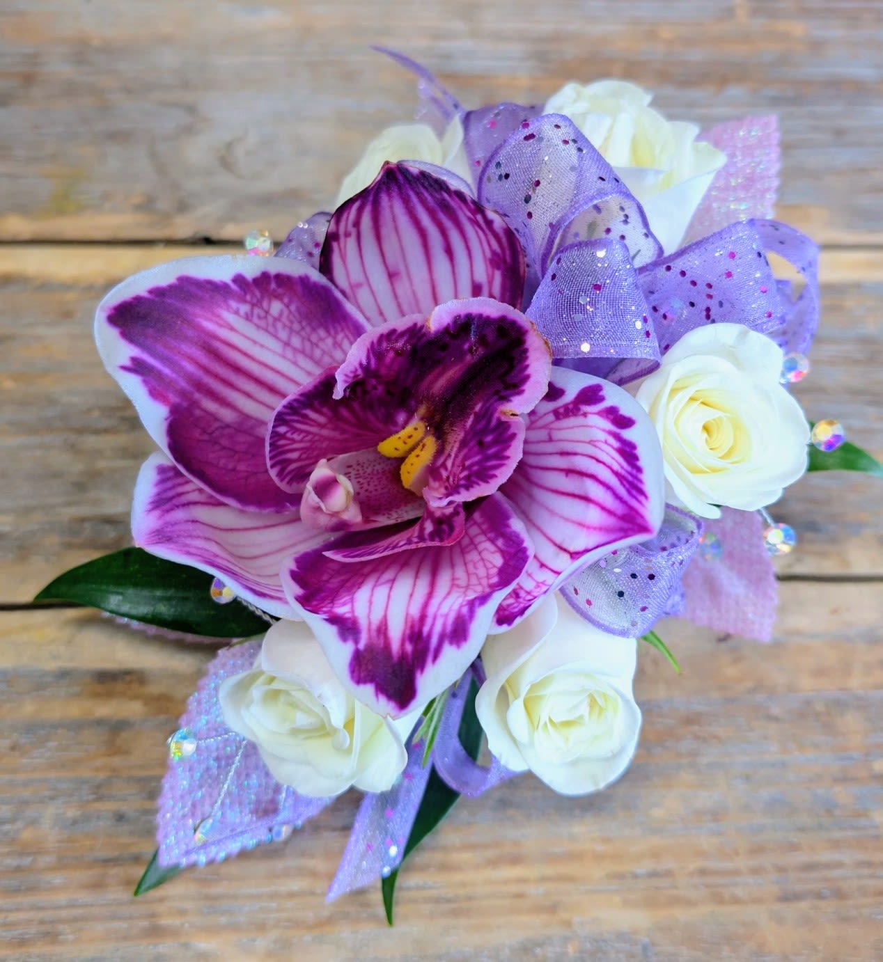 Orchid And Rose Corsage - Delicate, romantic, and unforgettable—our Orchid &amp; White Rose Corsage from Windmill Floral Expressions brings a perfect balance of elegance and color. A striking purple and magenta orchid takes center stage, its vibrant petals kissed with deep fuchsia veining, surrounded by soft white roses that symbolize timeless beauty. Accented with shimmering lavender ribbon, iridescent crystals, and lush greenery, this corsage captures both grace and sparkle in one exquisite design.  Perfect for proms, weddings, quinceañeras, or any formal celebration, this corsage is a wearable piece of art that elevates your look with fresh, handcrafted florals. Designed in Las Vegas with meticulous attention to detail, it’s an ideal choice for those who want to shine with sophistication and style.
