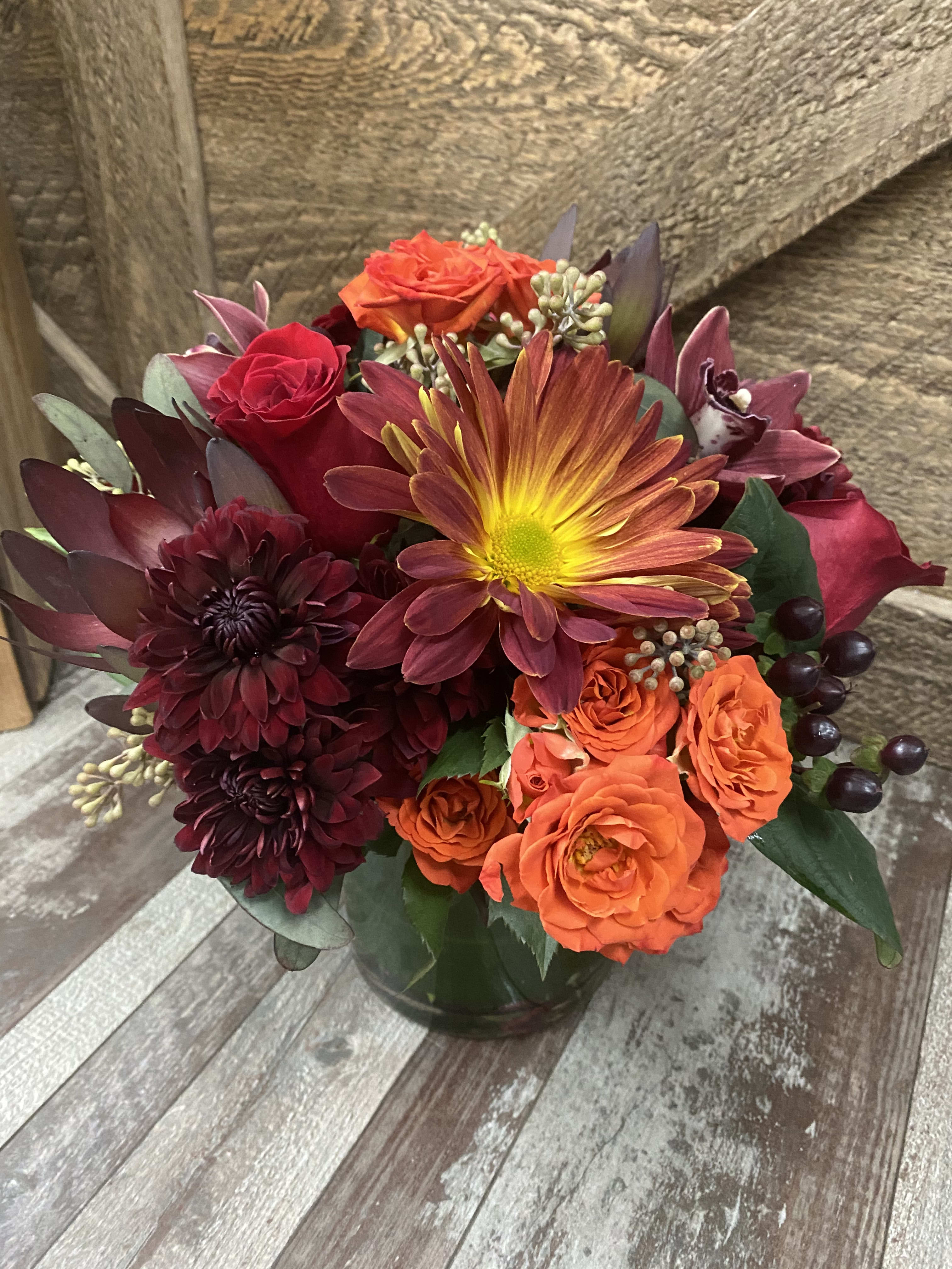 Fall is in the air - This lovely design includes orange and red roses, fall colored mums, berries, protea and eucalyptus. We arrange them in a clear glass leaf wrapped vase. A great addition to any holiday table. 