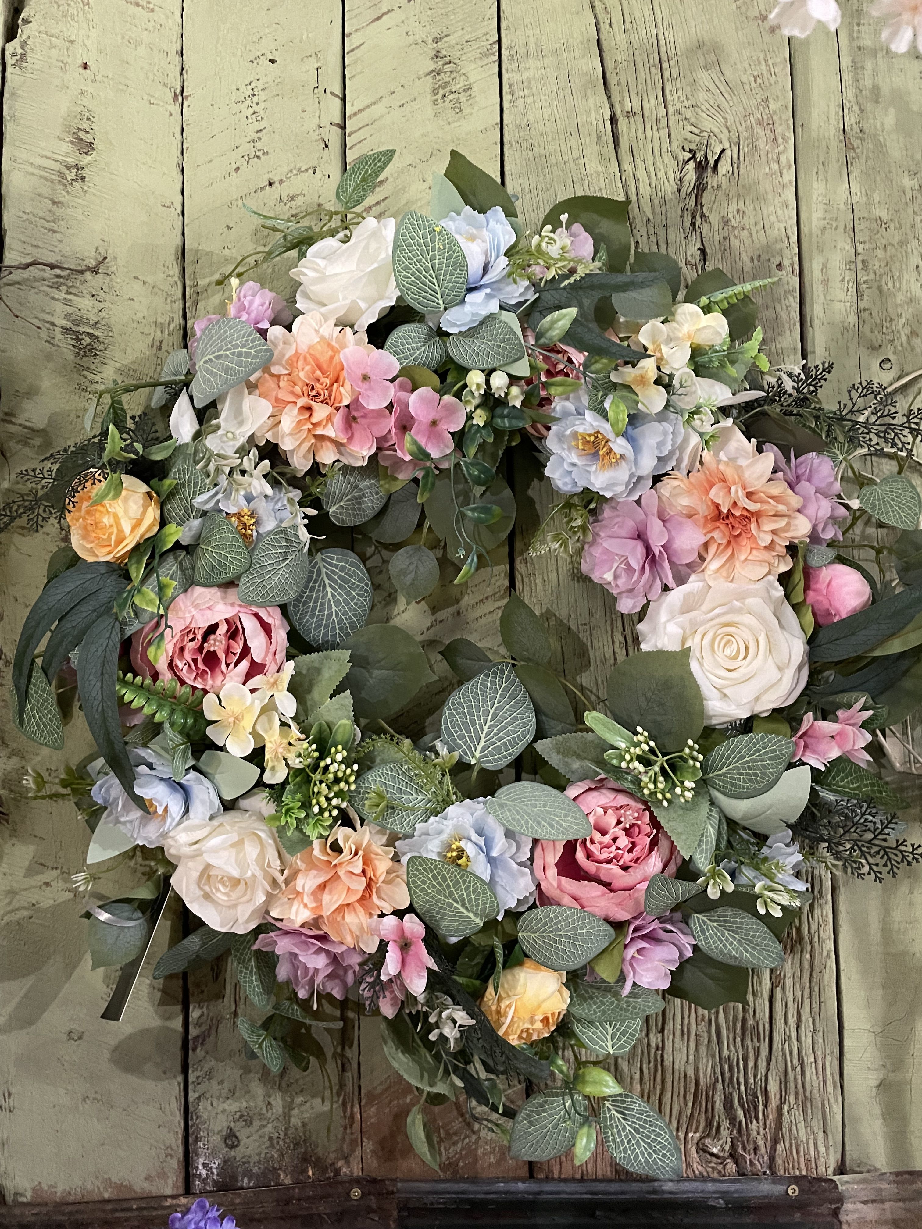 Lily Love Silk Grapevine Wreath - Bring a fun look indoors with a gorgeous grapevine accented in roses and mix. This wreath will look beautiful on a door or wall, and will brighten up the most dreary day. Mixed Greens with light pink lily florals.  Beautiful Grapevine wreaths are a stunning addition to any home. Hand made grapevine wreaths with high quality silk flowers. We create these wreaths in many different color schemes and price ranges. Perfect to send as sympathy gifts so that family can keep for years to come. Just pick your price and indicate what colors you prefer, we will create one Especially for you. Our stock changes frequently, so please call us @ 931-684-6863, if you have questions.  
