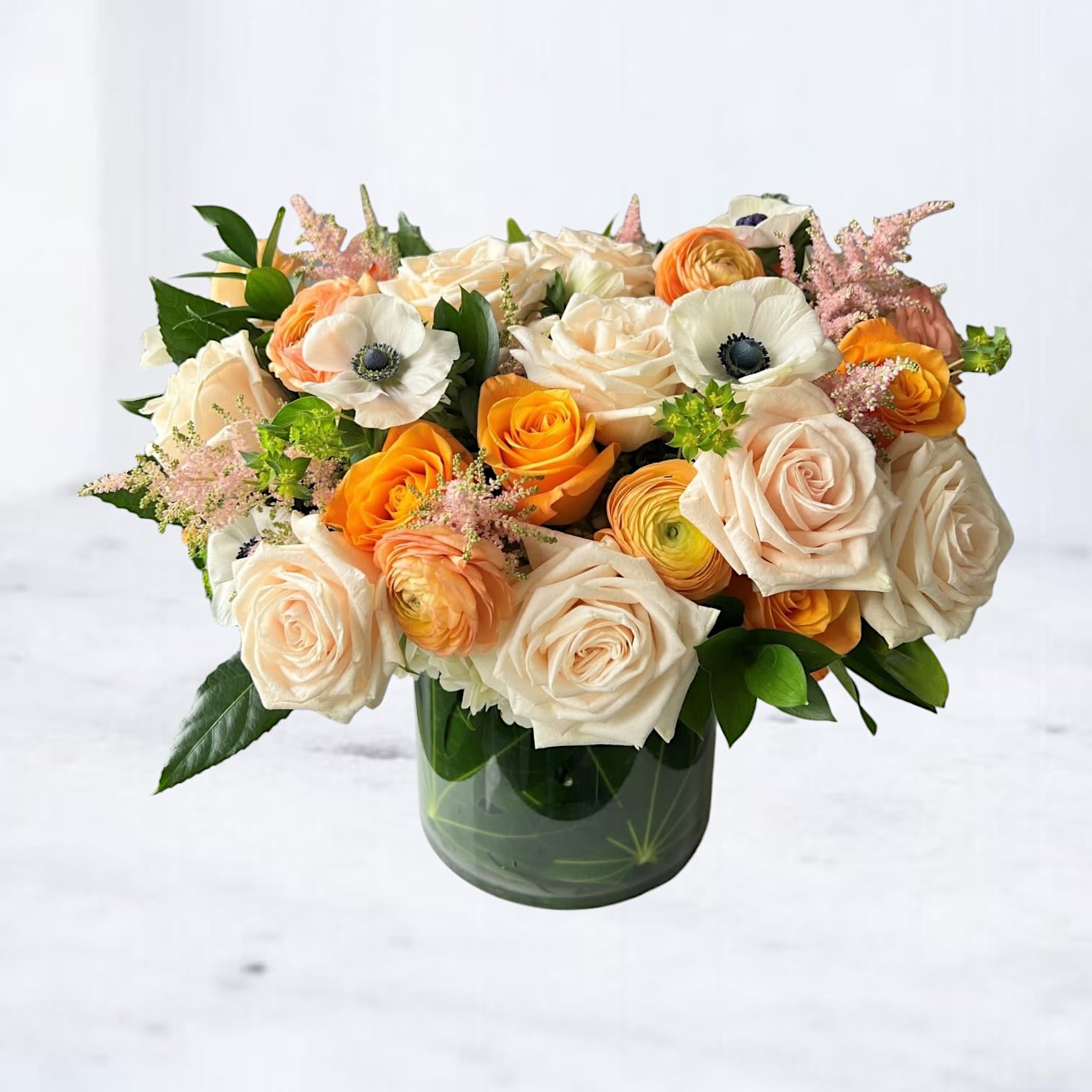 Vellum Glow Vellum Glow - Vellum Glow is a sophisticated floral arrangement designed to make a refined statement. Styled in a clear glass cylinder with a leaf wrap, this bouquet features peach and cream roses, vibrant orange ranunculus, white anemones with dark centers, pink astilbe, and fresh seasonal greenery for a layered, textural finish. Perfect for luxury gifting, anniversaries, or designer home décor, Vellum Glow delivers a rich, elevated color palette with soft movement and modern composition.