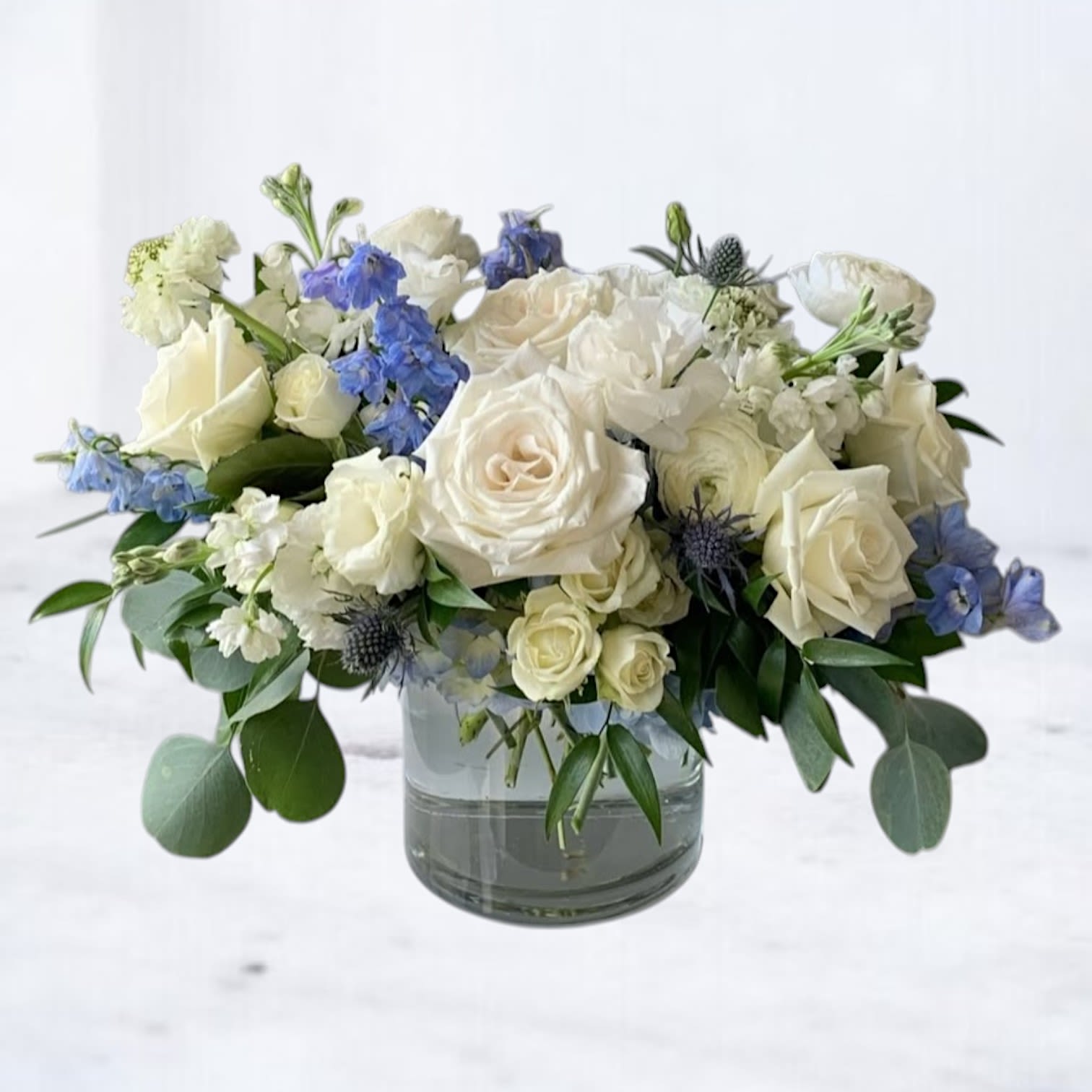 Ecliptic Aura Ecliptic Aura - A celestial composition of creamy white roses, soft lisianthus, icy blue delphinium, and striking thistle, Ecliptic Aura radiates elegance with a cool, refined palette. Designed in a clear glass cylinder vase, this arrangement evokes a sense of balance and calm—perfect for expressing sympathy, offering comfort, or simply bringing peace into any space.