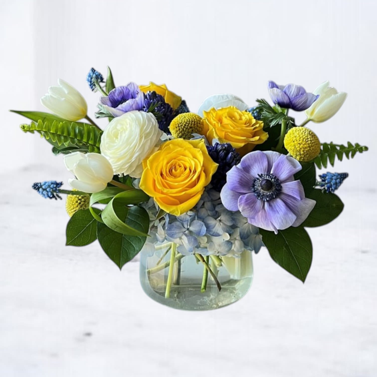 Sapphire Meadow Sapphire Meadow - Bright and unexpected, Sapphire Meadow brings together a striking palette of golden yellow roses, icy blue hydrangea, violet anemones, and creamy white tulips—accented with ranunculus, muscari, and craspedia for layered texture and personality. This vibrant floral design is perfect for celebrating birthdays, sending congratulations, or simply lifting someone’s spirits with something joyful and artful. Designed in a clear glass vase for a fresh, garden-inspired feel. Note: Muscari (grape hyacinth) is a seasonal bloom. When unavailable, we will substitute with a similar flower of equal or greater value to maintain the same vibrant aesthetic.