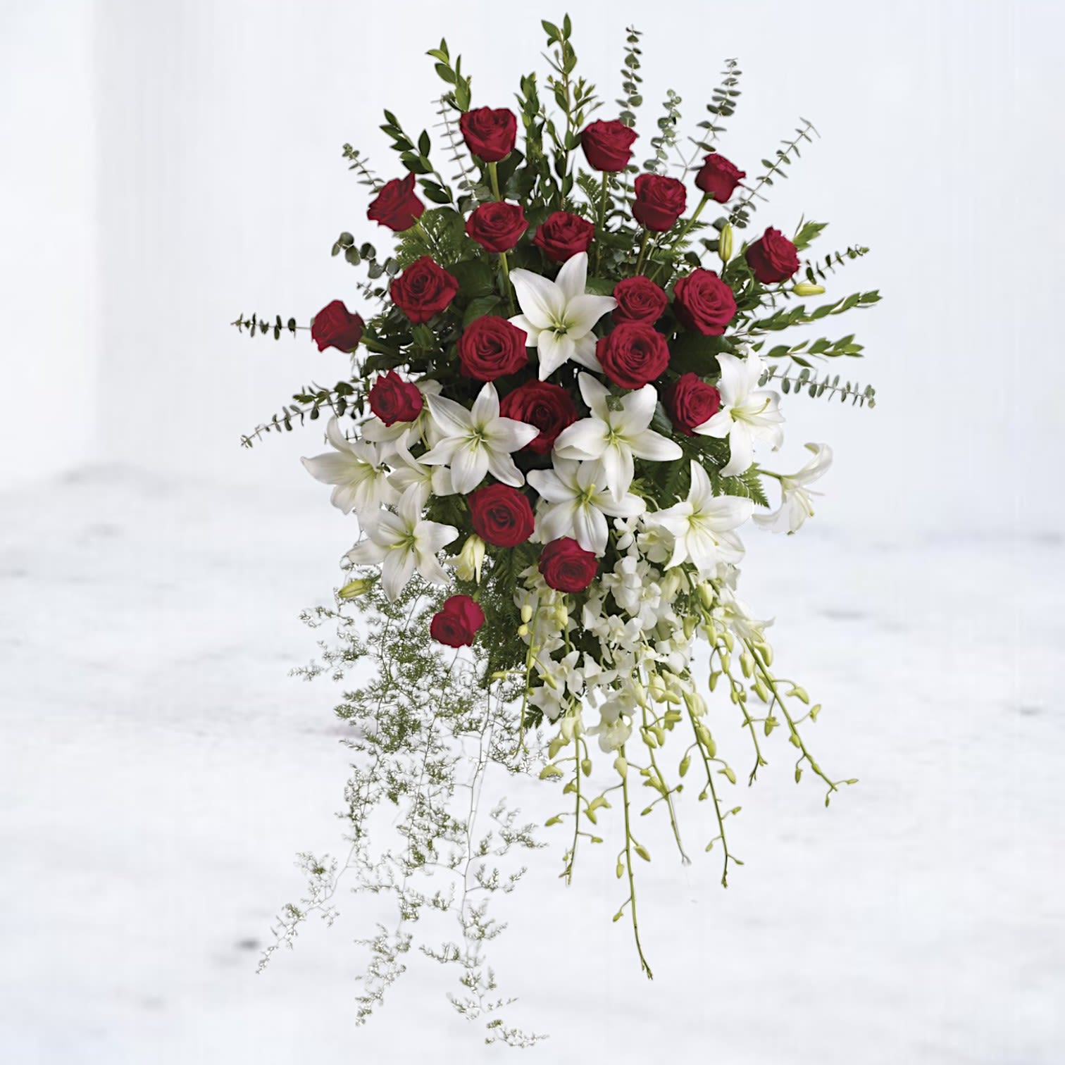 Sacred Tribute Standing Spray Sacred Tribute Standing Spray - Celebrate a life well-lived with the stunning Sacred Tribute. This elegant standing spray features a harmonious blend of lush red roses, pure white lilies, and cascading orchids, symbolizing love, peace, and everlasting remembrance. Accented with delicate greenery and filler florals, this design exudes sophistication and grace. Perfect for honoring a cherished loved one, this arrangement provides a heartfelt and timeless expression of sympathy. Placed on a sturdy easel, it offers a breathtaking tribute at funerals, memorials, or celebrations of life. Easels are included with all standing sprays for effortless display at services.