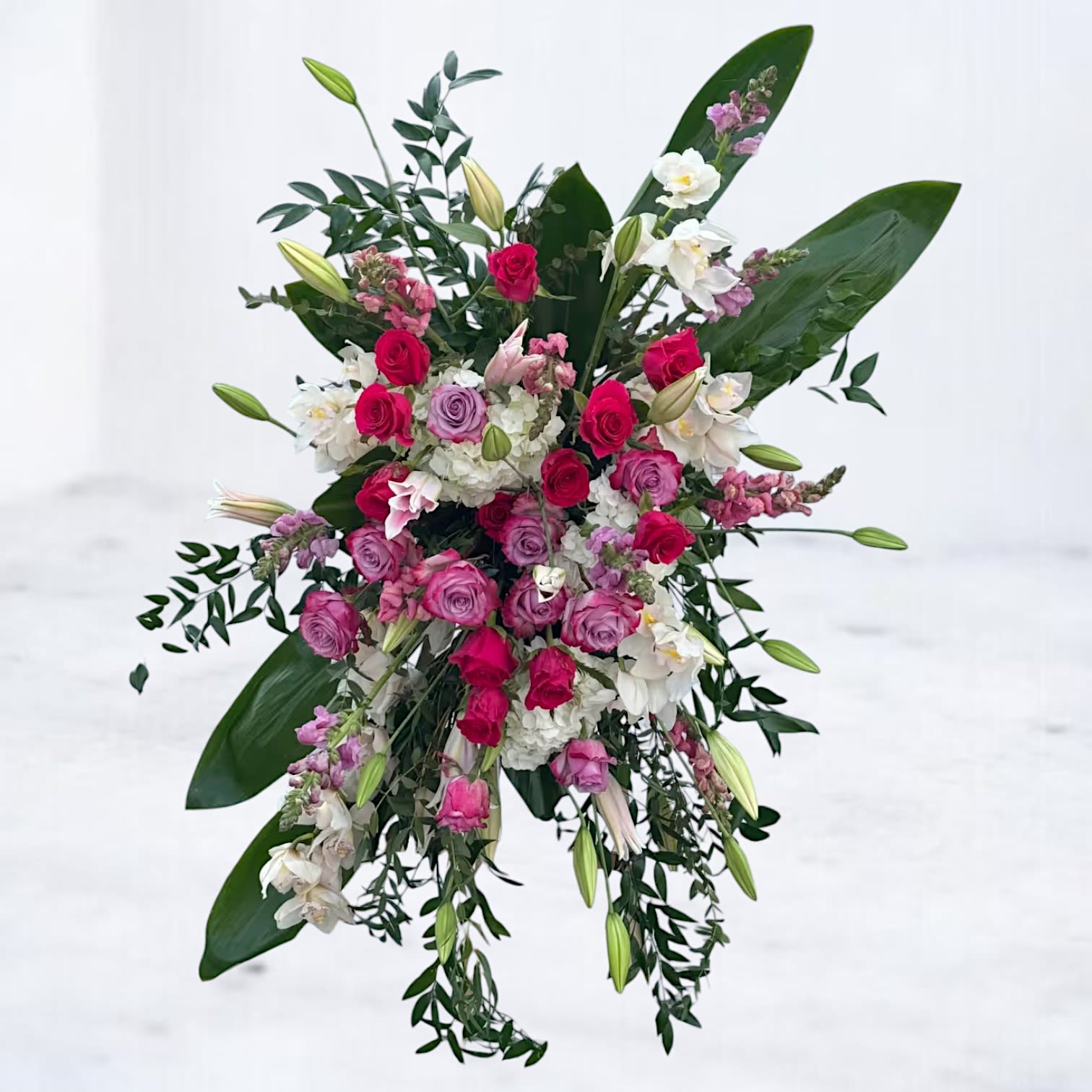Devotion Cascade Tribute Devotion Cascade Tribute - Elegant and expressive, the Devotion Cascade Tribute honors a life well lived with bold beauty and cascading grace. This standing spray features a striking composition of deep red and lavender roses, white lilies, soft pink snapdragons, and white hydrangea, accented with tropical foliage and flowing greenery. Designed to make a lasting impression at memorial or funeral services, this lush display offers comfort and remembrance in every bloom. Available for delivery to funeral homes and churches across Houston and surrounding areas.