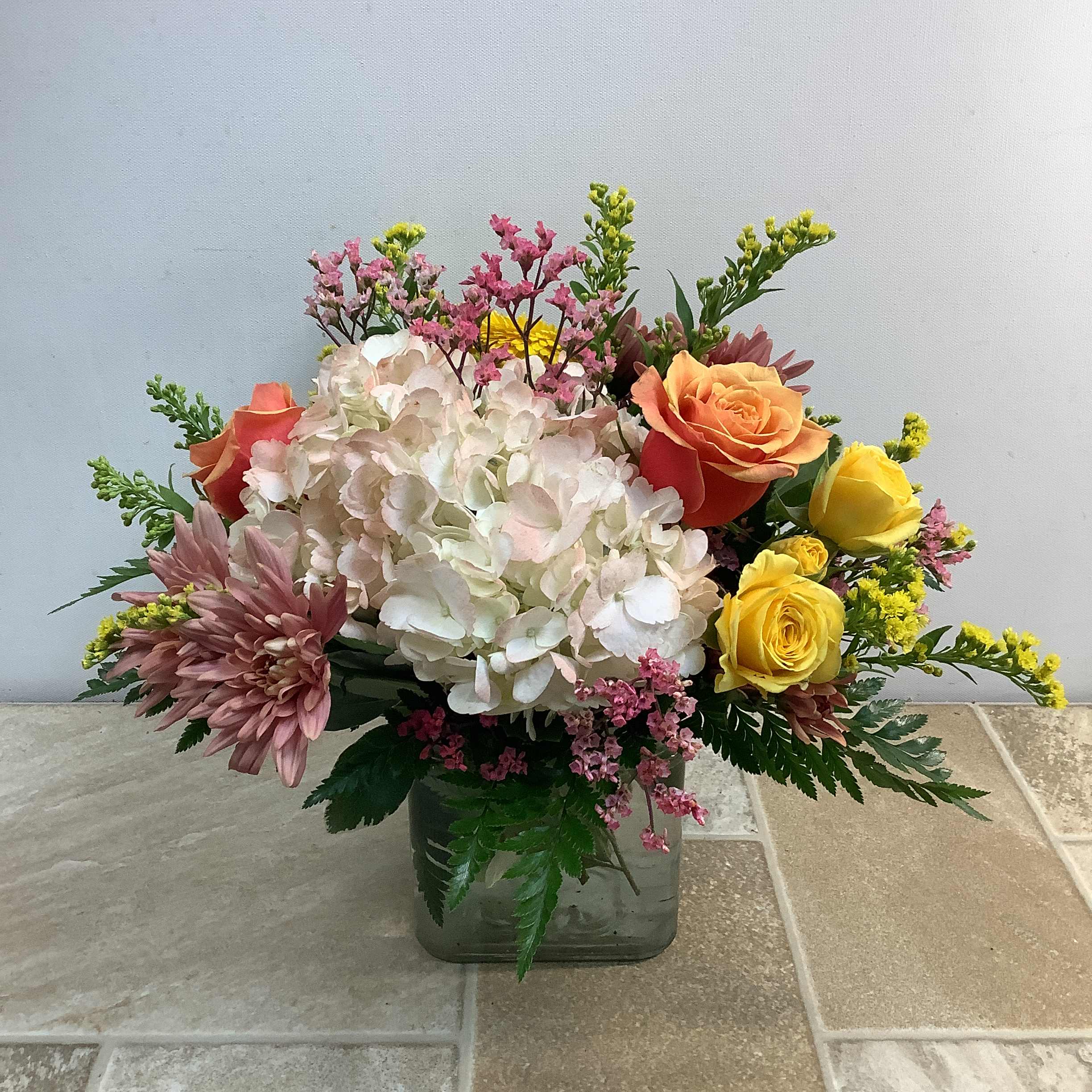 Blushing Beauty - The &quot;Blushing Beauty&quot; floral arrangement, presented in a glass vase, features a variety of flowers in a soft, romantic color palette. The central focus is a creamy blush-colored hydrangea, surrounded by lovely cherry brandy roses, bright yellow spray roses, a gerbera daisy, and peachy-pink mums. The blooms are accented by sprigs of accent flowers along with lush, leafy foliage. Approximate size: 15&quot; wide x 12&quot; high.  Primary flowers: hydrangea, rose, daisies, mum