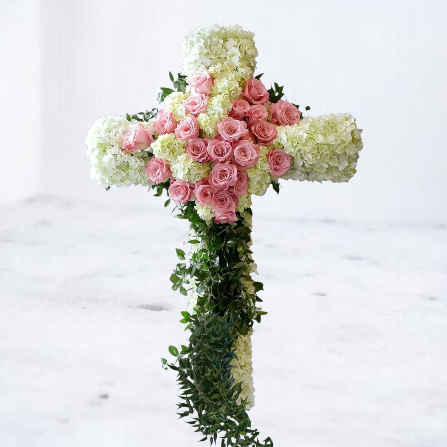 Sacred Devotion Funeral Cross - The Sacred Devotion Funeral Cross is a stunning tribute of faith and love, handcrafted with lush white hydrangea and soft pink roses, symbolizing purity, grace, and eternal devotion. Flowing ivy trails gently down the cross, adding a natural touch of serenity. Perfect for honoring a loved one’s memory, this floral cross brings a message of peace and comfort to any funeral, memorial, or celebration of life service.   All floral crosses come with easels included for seamless presentation at memorials or services. 