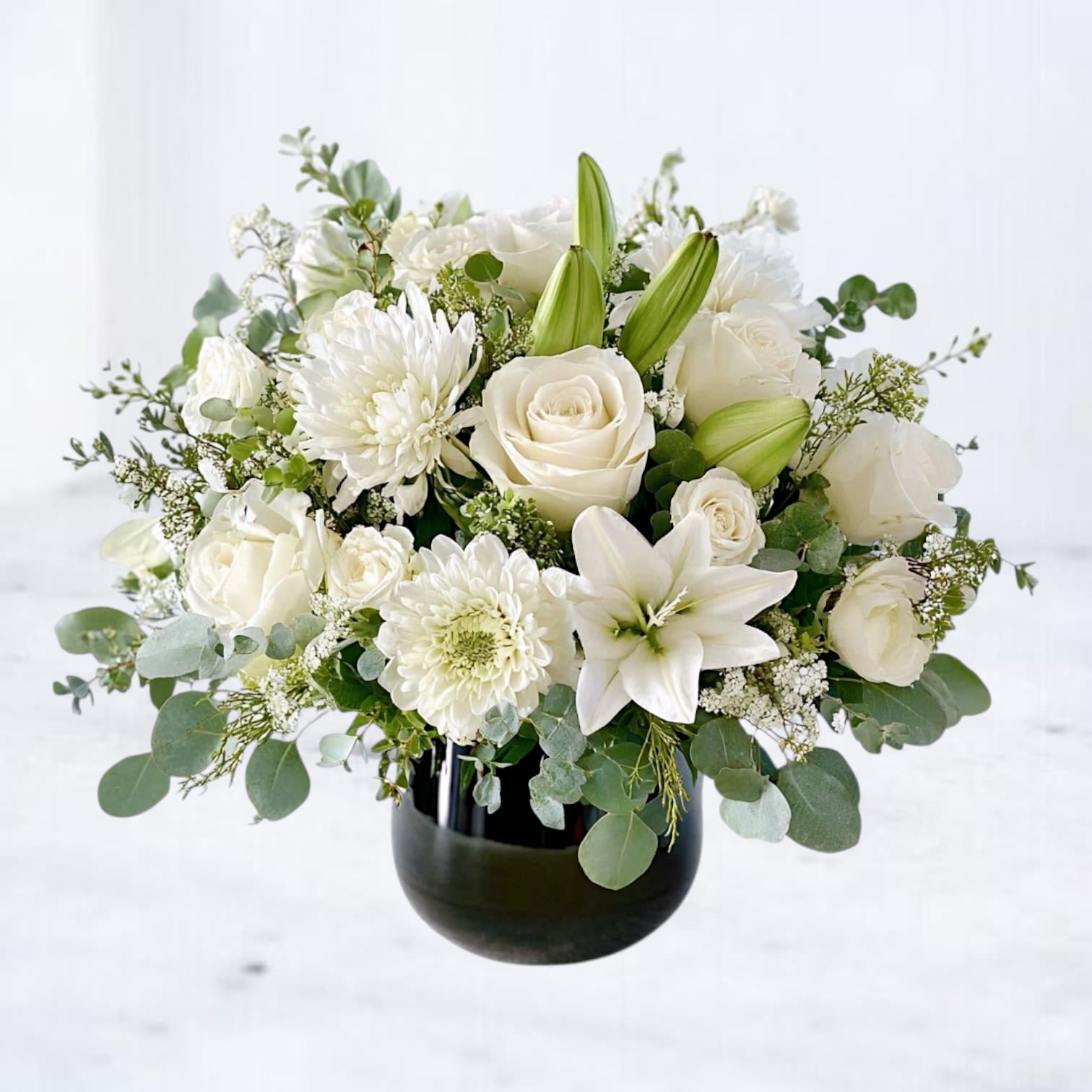 Pure Remembrance Pure Remembrance - The Pure Remembrance bouquet is a timeless expression of peace and comfort, featuring pure white roses, lilies, and seasonal blooms accented with soft greenery. Designed in a sleek black vase, this arrangement carries a modern yet classic presence, offering solace during times of loss. Perfect for funeral services, sympathy gestures, or sending condolences to the home, this all-white bouquet symbolizes purity, reverence, and everlasting love. Its refined design brings quiet beauty and heartfelt comfort to those who need it most. Available for delivery in Magnolia, The Woodlands, Spring, Montgomery, and surrounding Houston areas.