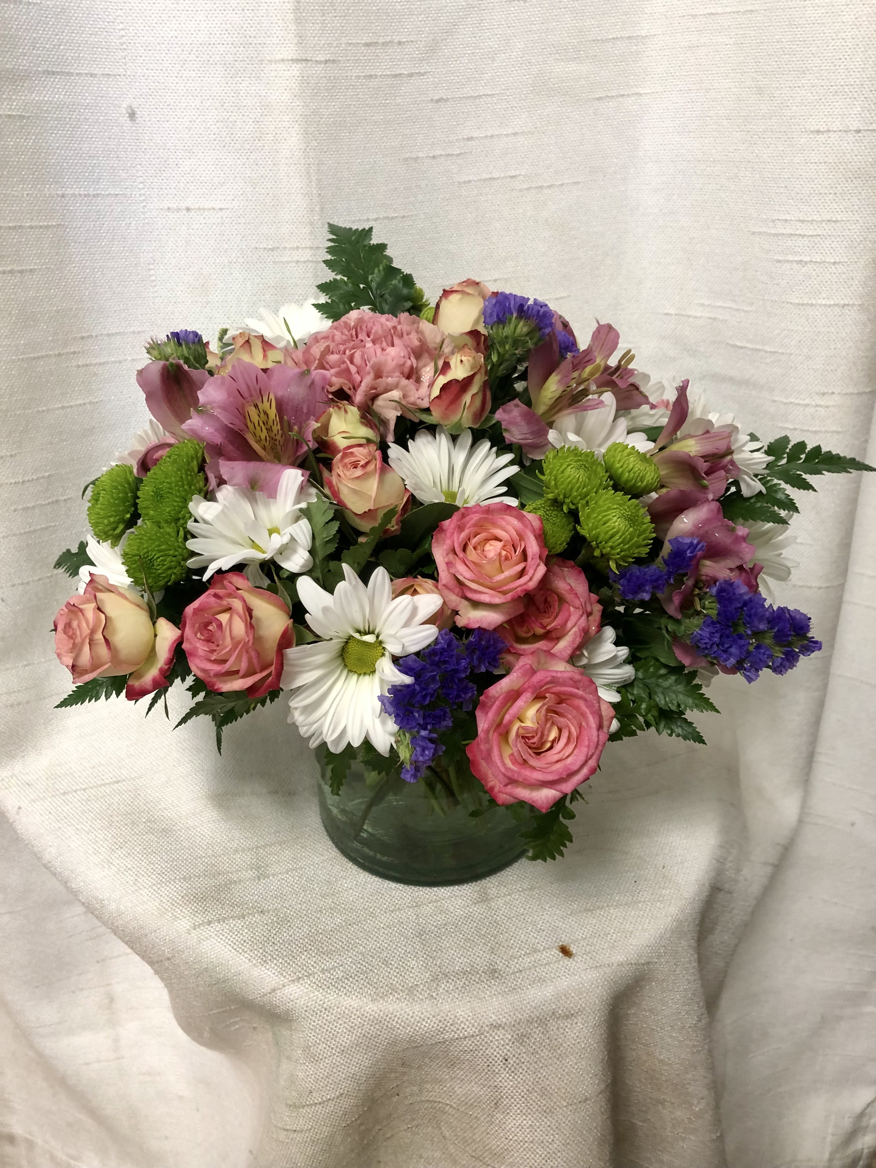 Berry Bliss (BR) - Introducing our Berry Bliss Floral Arrangement, a vibrant and charming display of nature's beauty. This delightful arrangement features a mix of purple statice, green button poms, pink spray roses, white daisies, pink alstroemeria, waxflower and pink carnations, all lovingly arranged in a clear glass 5x5&quot; cylinder vase.   