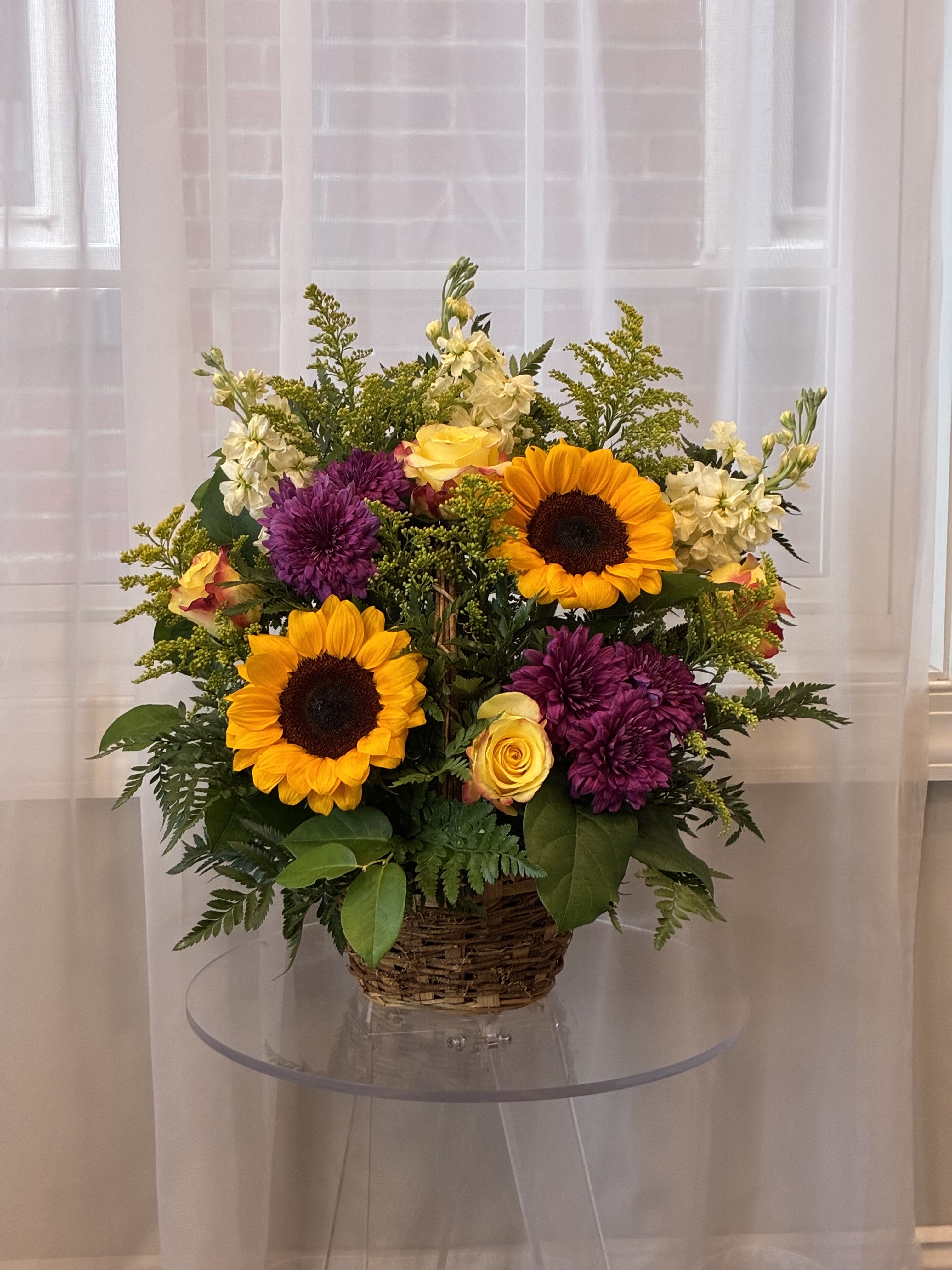 Autumn Harvest Basket - Bring the warmth and richness of the season into any space with our Autumn Harvest Basket. This charming arrangement overflows with the golden glow of sunflowers, the vibrant elegance of orange roses, and the deep jewel tones of purple chrysanthemums. Accented with solidego, ivory stock, and seasonal foliage, this handwoven basket captures the cozy beauty of fall.  Please note, the picture under the standard size is actually the deluxe size!