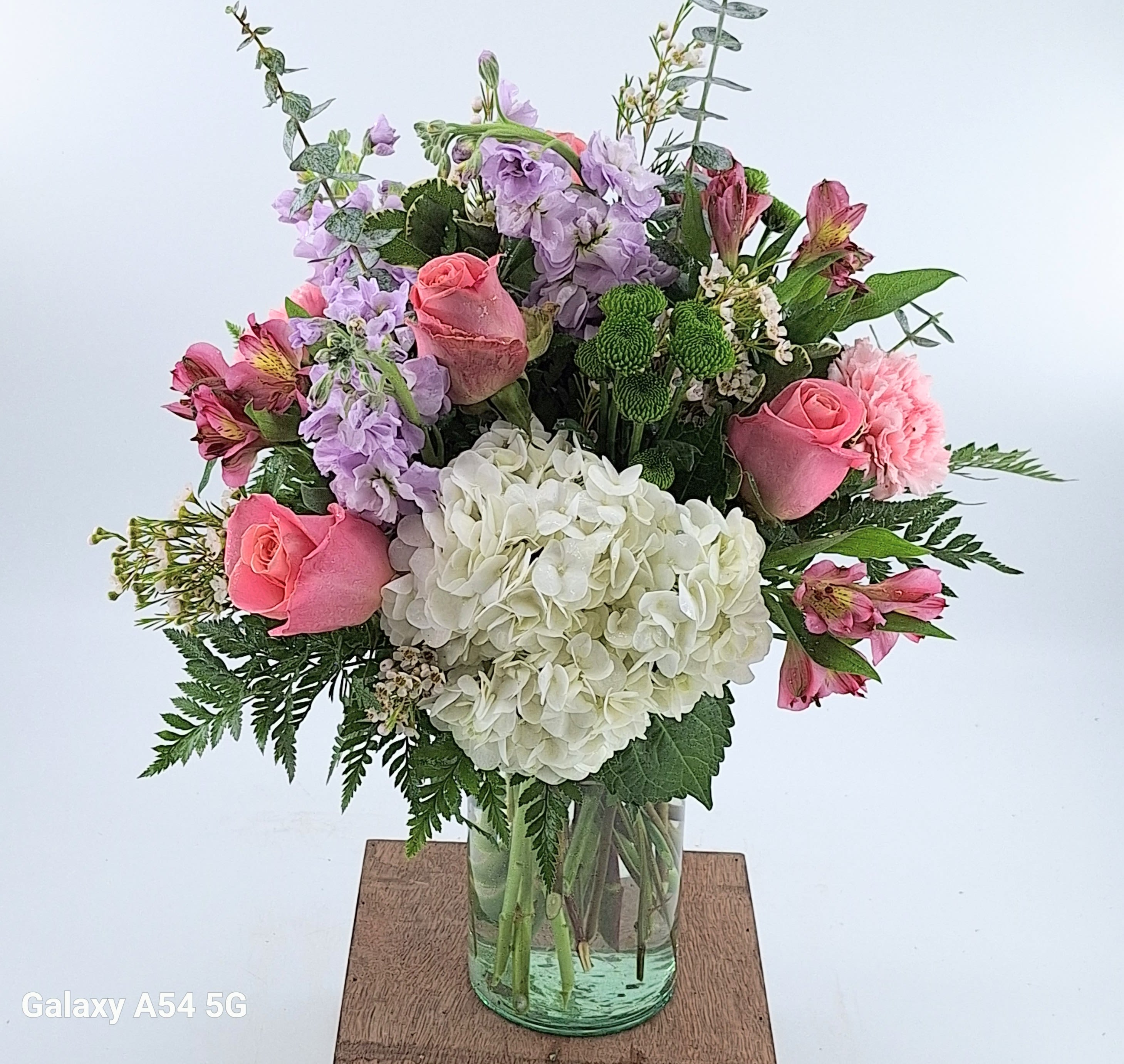 The April (BR) - This enchanting bouquet features an exquisite combination of pink roses, white hydrangeas, lavender stock, white carnations, pink alstroemeria, green button poms, and lush greenery, perfectly arranged in a traditional glass vase.  