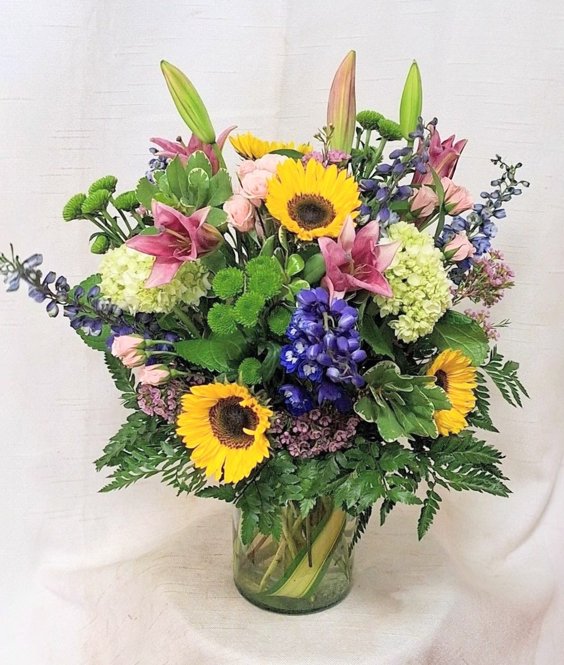 Summer Breeze (BR) - Brighten any space with this vibrant flower arrangement featuring pink lilies, hybrid blue delphinium, sunny sunflowers, delicate pink spray roses, green button poms, and fragrant pink waxflower, elegantly displayed in a leaf-lined glass vase for a fresh, natural touch. 