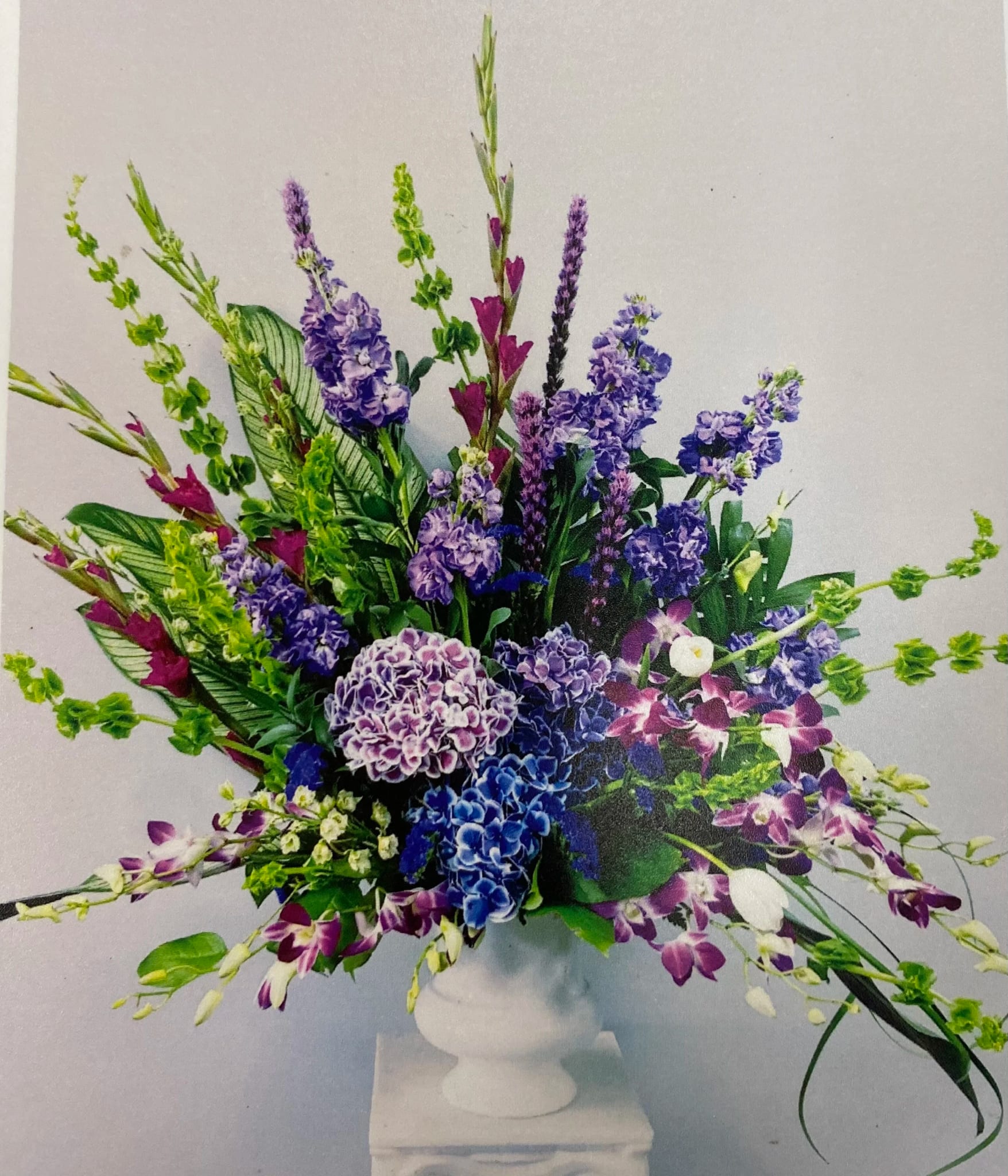 Ceremony Arrangement CV-22 Ceremony Arrangement CV-22 - A striking expression of reverence, this arrangement is filled with layers of deep purples and vibrant greens. Featuring stock, hydrangea, dendrobium orchids, liatris, and bells of Ireland, this design evokes a sense of dignity and reflection, making it a graceful tribute for a memorial or service.