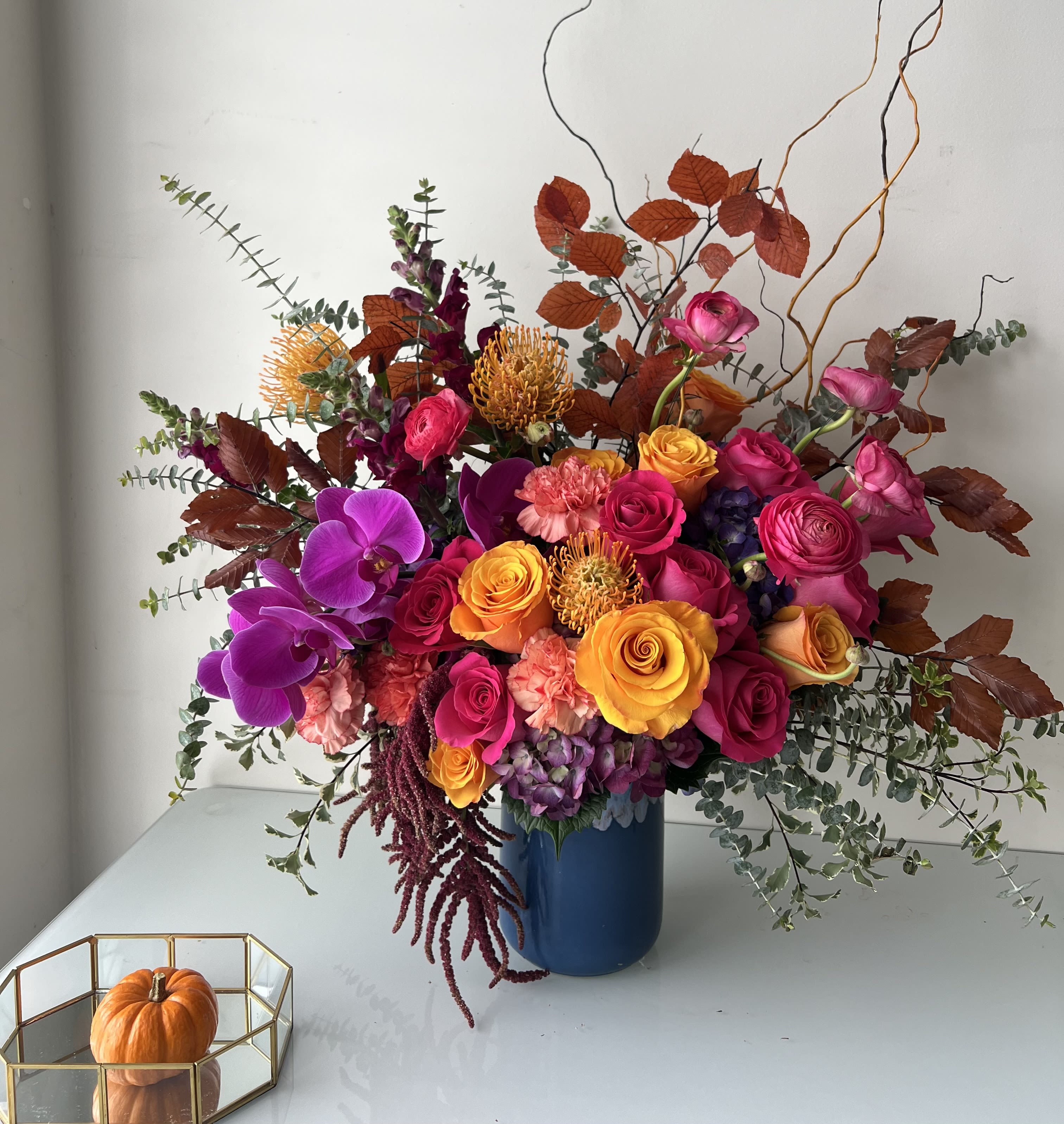 A California Autumn - We all know those sunny California days in Autumn. A bright and upbeat take on Autumn.   Flowers: orange roses, orange pincushions, pink floyd roses, hot pink ranunculus, purple jewel orchids, royal purple variegated hydrangea, coral carnations, burgundy hanging Amaranthus, wine snapdragon, burnt orange autumn leaves, baby blue eucalyptus, and curly willow.   Arranged in our Blue Portugal Pot