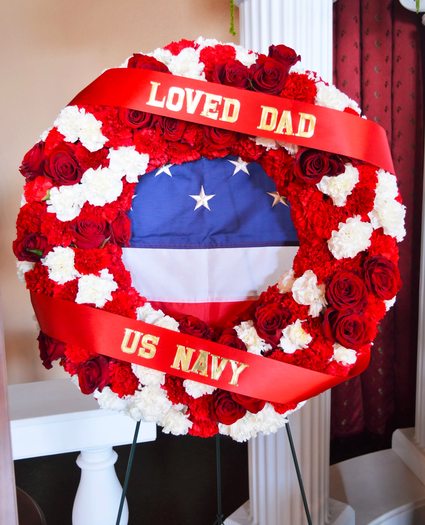 Wreath WR-06 Wreath WR-06 - Honor a life of service and devotion with this patriotic wreath, featuring a bold mix of red roses, white carnations, and vibrant red carnations in a classic circular design. The American flag is nestled within the center, symbolizing courage and sacrifice. This tribute is especially meaningful for military funerals or veterans' services throughout Goodyear, Buckeye, and Litchfield Park. Easel stand included; ribbon lettering available for an additional charge.