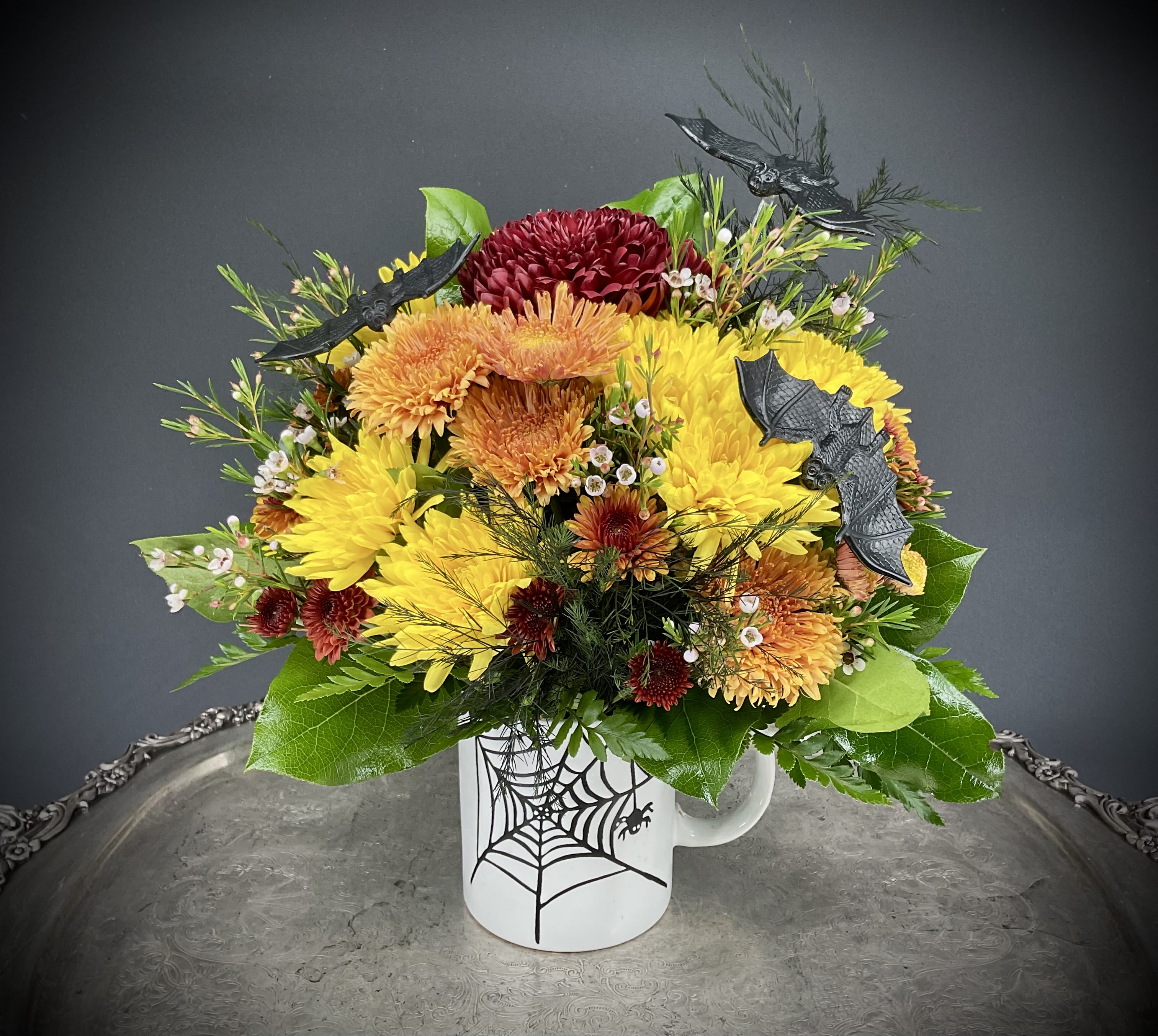 Halloween Spooktacular - A whimsical floral arrangement bursts from a keepsake (and reusable) coffee mug, featuring vibrant orange, red, and yellow blooms. A lifelike spider perches playfully on the front in its web, while bats hover above, adding a touch of eerie charm. Perfect for spooky autumn décor with a delightful blend of beauty and Halloween mischief.