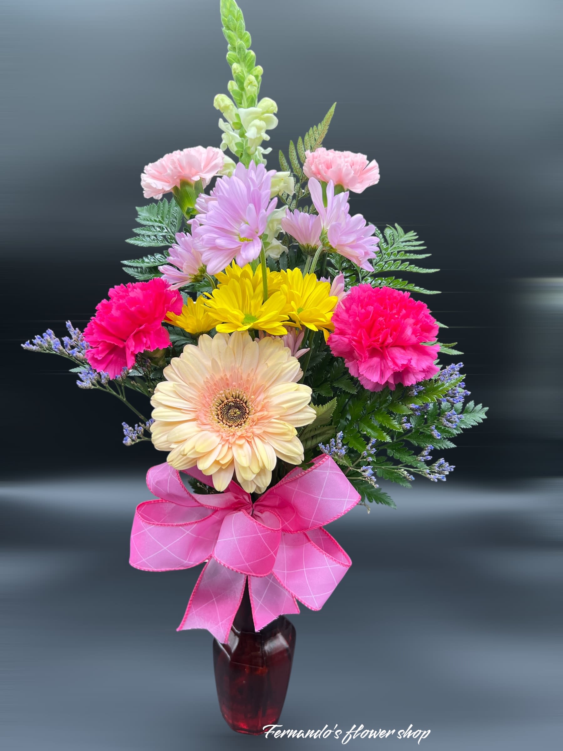 Just Reminding you! - This cheerful and vibrant floral arrangement is a delightful blend of colors and textures, perfect for bringing joy to any occasion. Featuring a soft peach gerbera daisy as the centerpiece, the bouquet is surrounded by bright pink and yellow carnations, delicate lavender daisies, and pastel pink snapdragons. Accents of lush greenery and hints of purple filler flowers enhance the natural beauty of the design. Tied together with a charming pink ribbon bow and displayed in a sleek red vase, this arrangement from “Fernando’s Flower Shop” exudes warmth, happiness, and positivity. Ideal for celebrations, thank-you gestures, or simply brightening someone’s day.