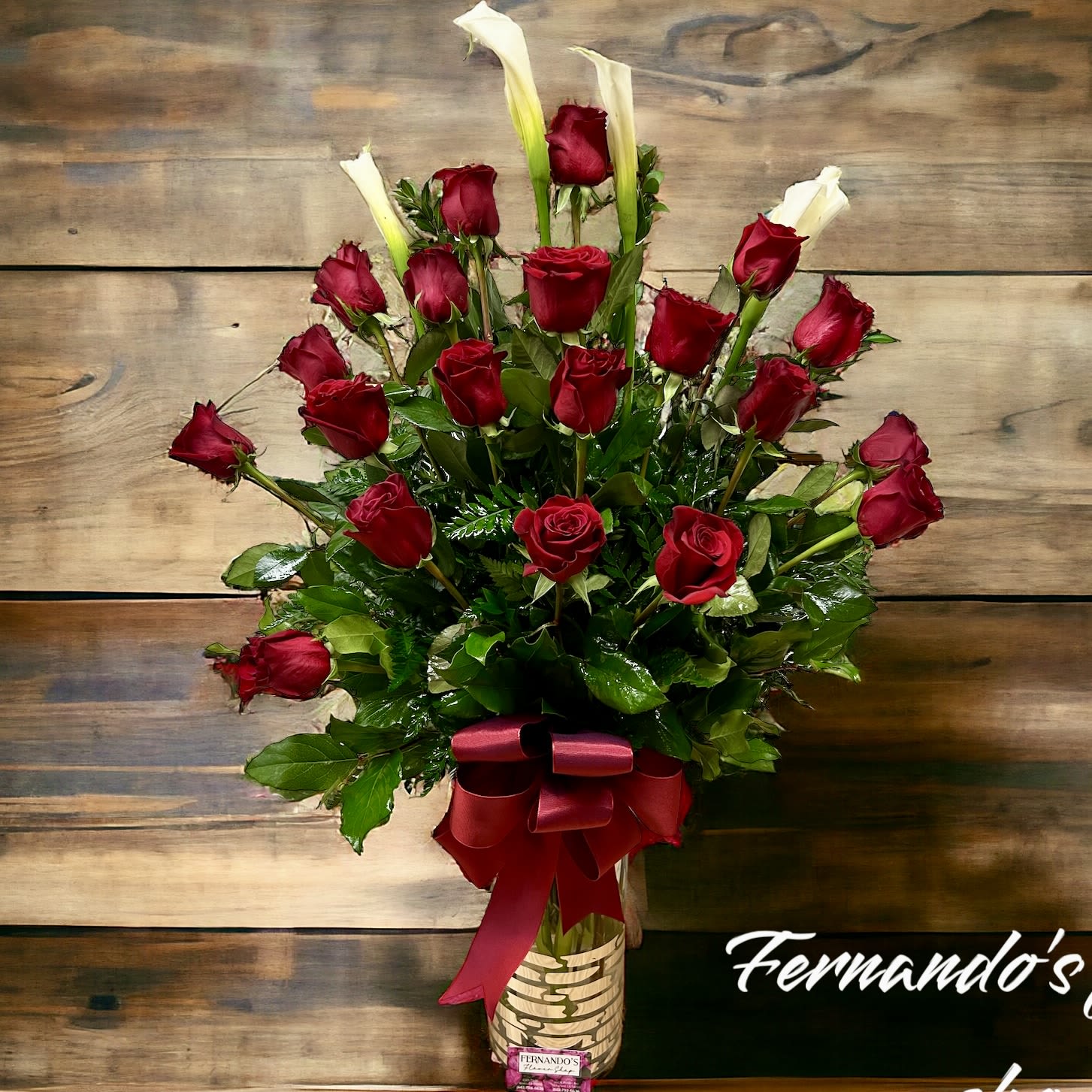 Blooming Grace - The image shows an elegant floral arrangement consisting of deep red roses and white calla lilies. The flowers are arranged in a fan-like shape, surrounded by lush green foliage, and placed in a decorative vase. A red ribbon bow adorns the vase, adding a sophisticated touch. The display is set against a rustic wooden background on a simple wooden table, giving it a warm, natural feel. The text “Fernando’s flower shop” is displayed on the lower right corner.