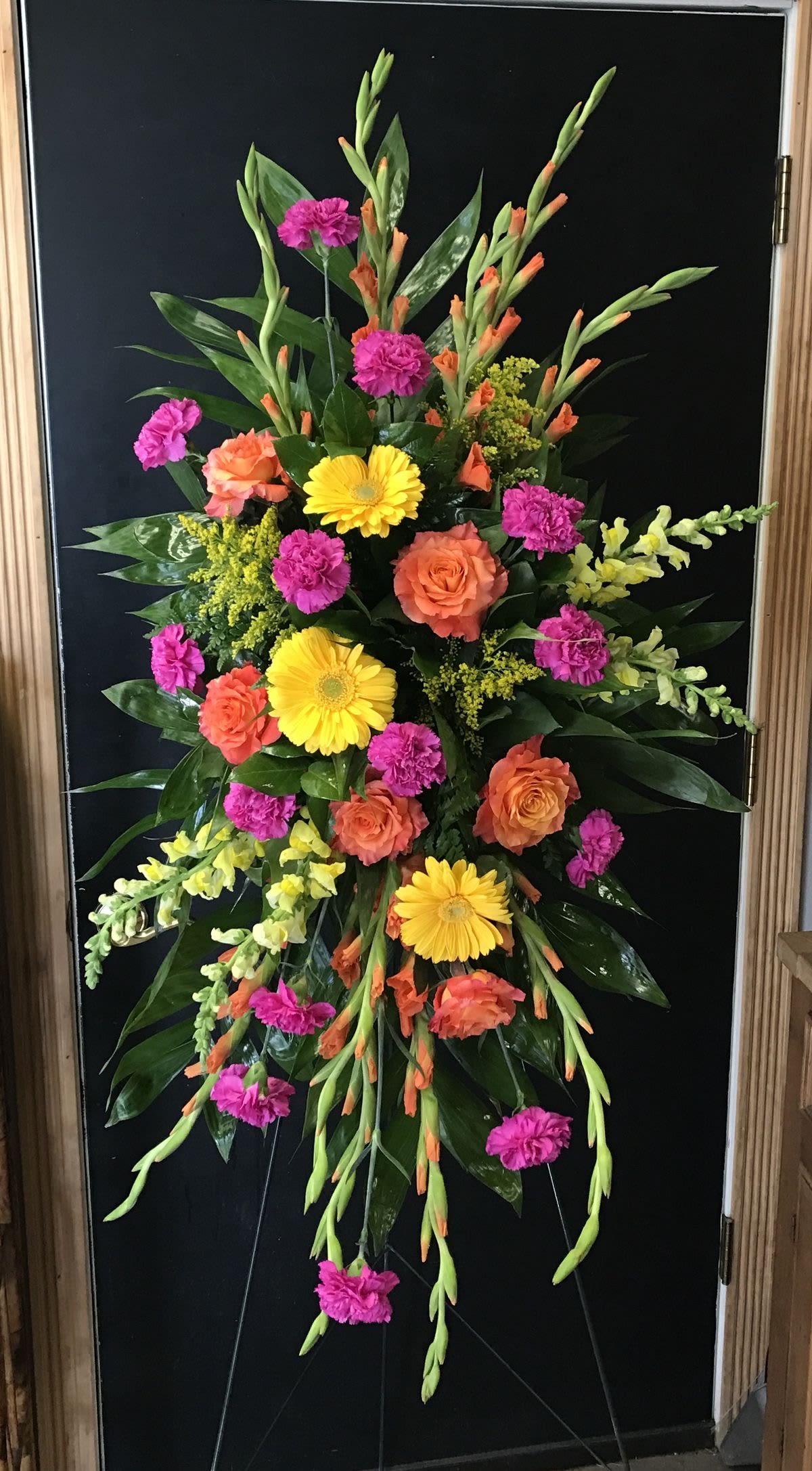 Happiness  - This is a vibrant and elegant standing floral arrangement featuring a mix of bright yellow gerbera daisies, orange roses, pink carnations, and yellow snapdragons. The display is accented with lush greenery and orange gladiolus, arranged in a cascading design. The bold colors and flowing composition make it a striking and cheerful floral tribute.