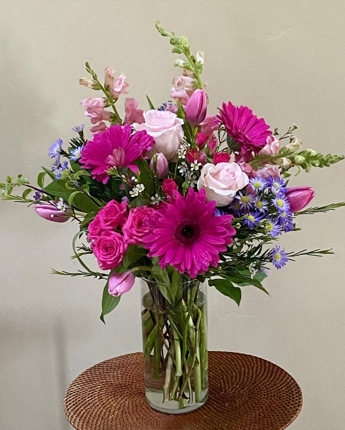 Ever After - This is a beautifully arranged floral bouquet in a clear glass vase, featuring a mix of pink and purple blooms. The arrangement includes vibrant pink gerbera daisies, soft pink roses, delicate snapdragons, tulips, and small purple asters, complemented by lush greenery and filler flowers. The combination of shades of pink, purple, and white creates a romantic and elegant display, perfect for adding a cheerful and sophisticated touch to any space.