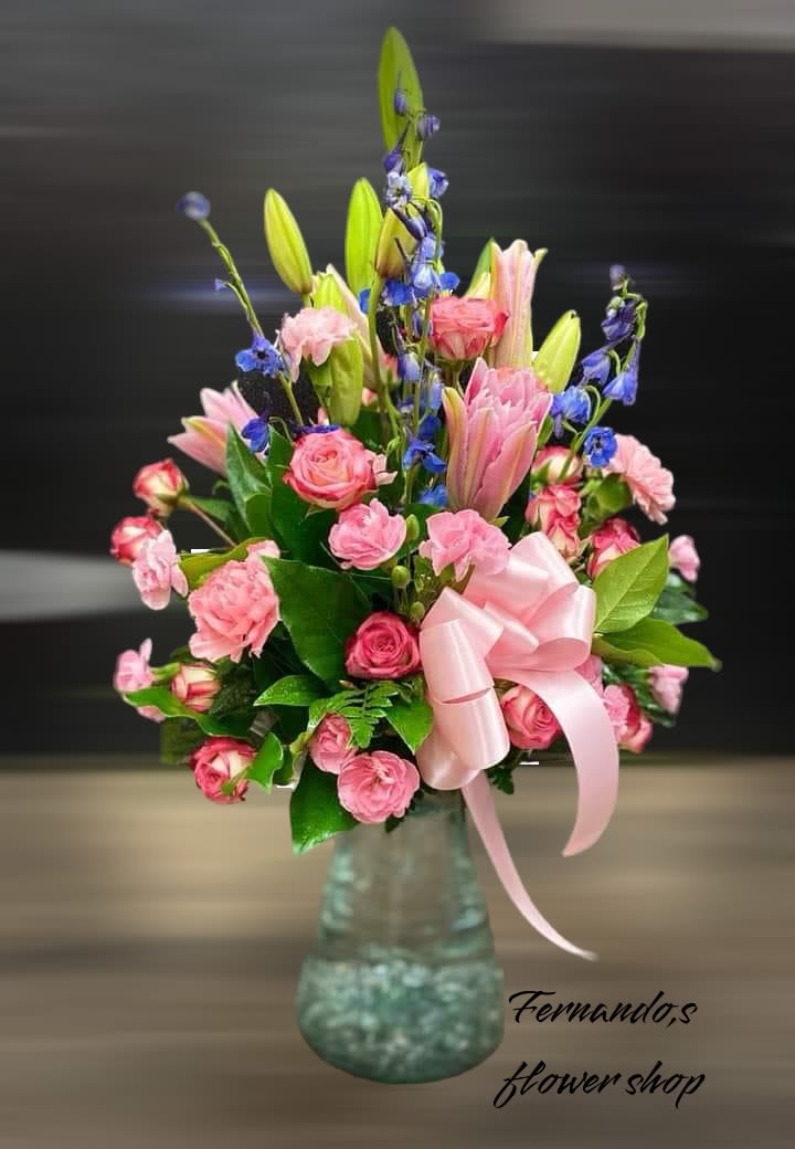 Pinks &amp; Blues - This elegant floral arrangement features soft pink roses, pink lilies, and vibrant purple larkspur, creating a delicate yet striking display. Lush green foliage adds contrast, while a pale pink ribbon bow enhances the arrangement’s romantic charm. Presented in a textured glass vase, this bouquet is perfect for expressing love, celebrating special occasions, or offering a thoughtful gesture. The bottom of the image features the text “Fernando’s Flower Shop,” indicating the florist.