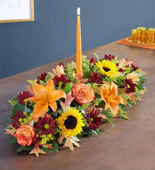  Thanksgiving Centerpiece with Candles - A Thanksgiving centerpiece filled with our beautiful flowers in fall-themed colors. All-around arrangement with autumn-colored roses, sunflowers, red daisy poms, orange Asiatic lilies, mini carnations and solidago; accented with assorted greenery and dried oak leaves