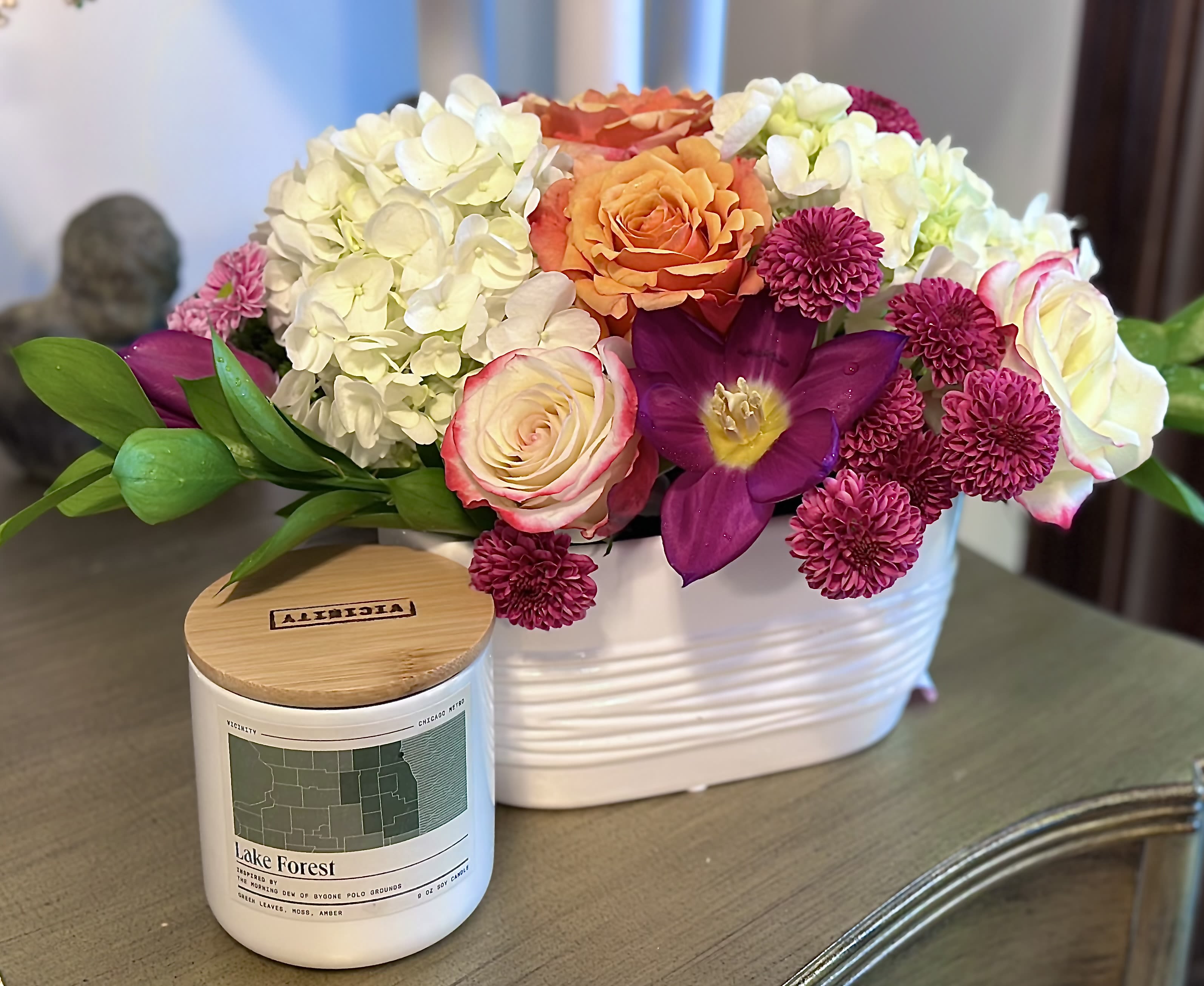 Garden Bouquet &amp; Highland Park Candle - What better way to show a Northshore resident you care than with a gorgeous garden bouquet arrangement and a candle by locally made Vicinity Candles!   Petite Bouquet: A 6&quot; W x 6&quot; H collection of seasonal blooms in a clear glass vase. Please note that colors may not match the picture because we source new flowers and colors weekly. Be assured that your arrangement will feature colorful and lush blooms.   The Highland Park Candle: Inspired by a cozy, blanketed sunset picnic at Ravinia, with notes of bergamot, amber, and gardenia.  Located in the heart of the North Shore, Highland Park was named by former Chicago Mayor Walter Gurnee for its elevated and picturesque, park-like setting. In the 1850s, the railroad brought Chicago’s upper echelon, who envisioned a high-end amusement park called Ravinia Park. Ravinia instead grew into a world-renowned music venue attracting over 400,000 guests to its expansive lawn each summer. Ravinia, along with the area’s peaceful setting and tightknit community nestled just outside Chicago, make Highland Park a wonderful place to live.  Source: Encyclopedia of Chicago.  Vicinity Candles is a local Chicago business that crafts handmade soy candles inspired by Chicagoland's unique neighborhoods and towns.  9 oz soy candle | Burn time: 45-50 hours | Packaged in a branded gift box