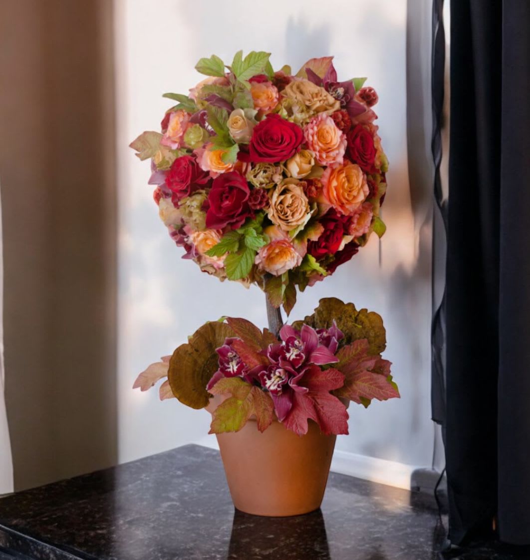 Woodland Fall Woodland Fall - A miniature autumn tree arrangement featuring a harmonious fusion of fall hues. This composition showcases a spherical cluster of roses, with enchanting shades of red, peach, and nude, adorned with intertwined autumn foliage. Nestled within a rustic terra cotta pot, the base is further embellished cymbidium orchids, and additional fall foliage. Standing at a height of 24 inches, this tree-like display is crowned by a resplendent rose ball measuring 12 inches in diameter.