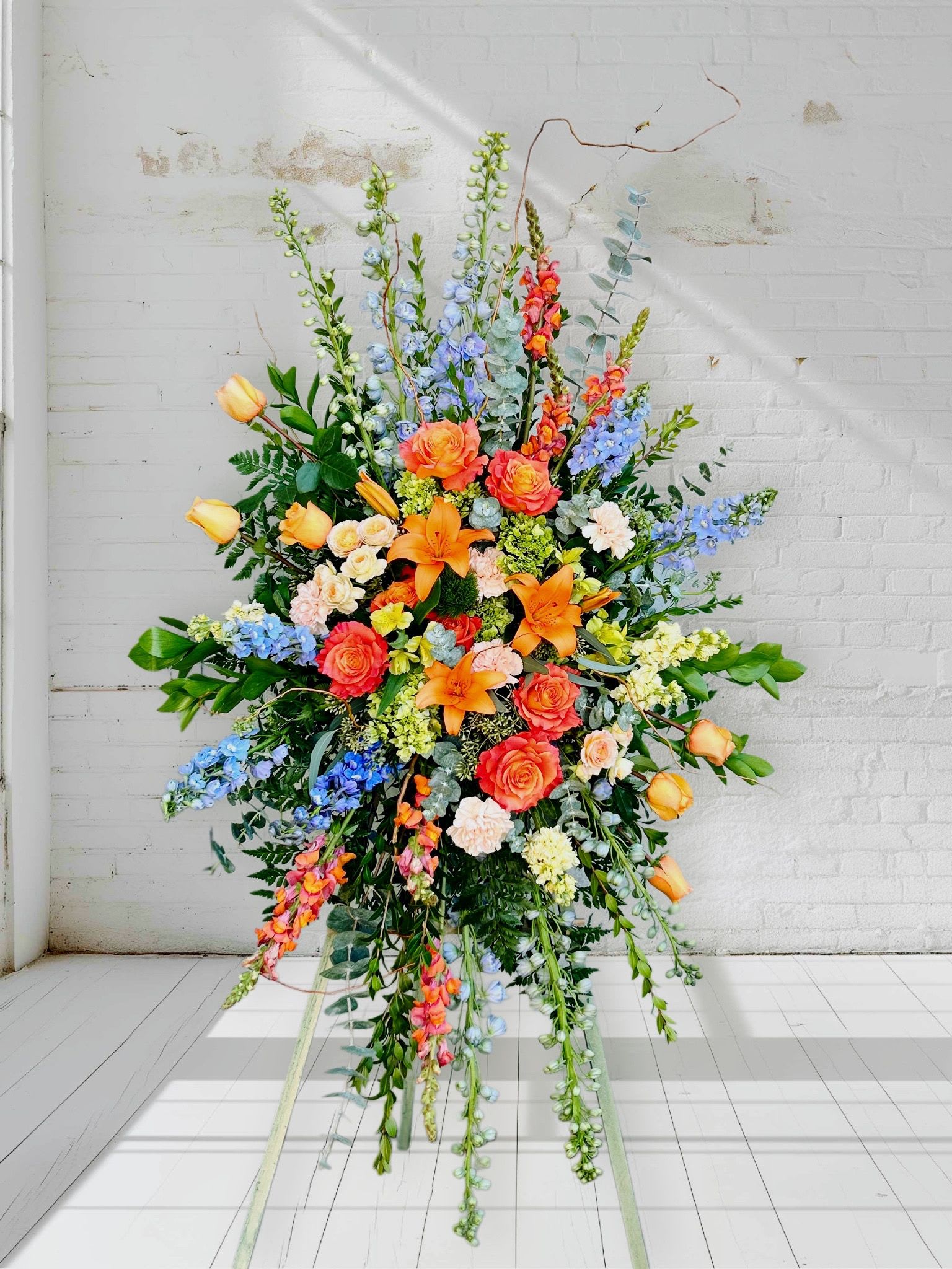 Bright Horizon Standing Spray - A radiant standing spray designed to bring comfort and hope in times of sorrow. Bright Horizon blends the warmth of orange lilies and roses with the soothing tones of blue delphinium, soft peach blooms, and touches of yellow. Cascading greenery and elegant textures give the design a sense of movement—like sunlight breaking through the sky after a storm.  This arrangement honors a beautiful life with both grace and vibrancy, offering a heartfelt message of remembrance and the promise of brighter days ahead.
