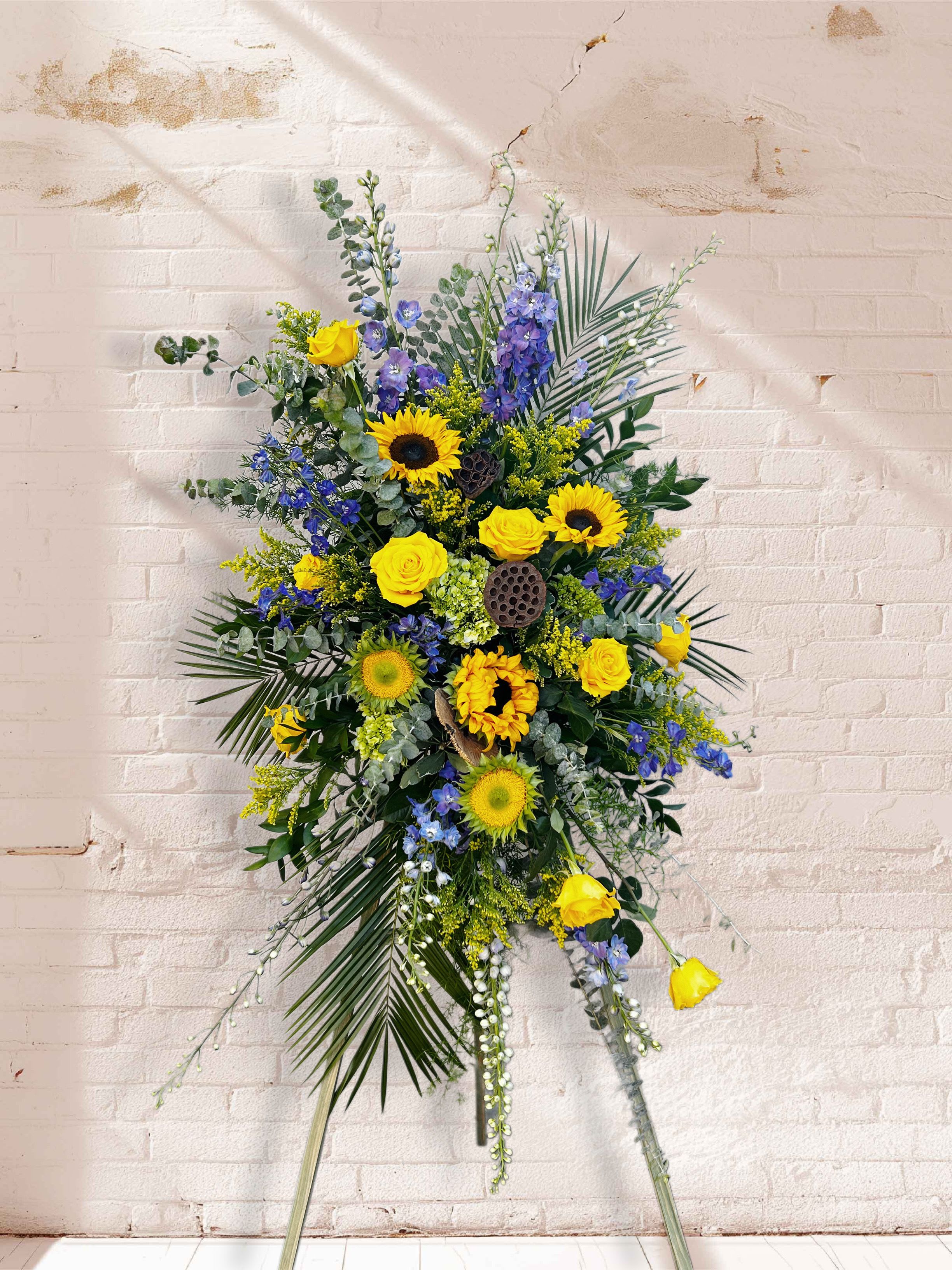 Ray of Hope Standing Spray - Bring warmth and comfort to a time of loss with our Ray of Hope standing spray. Golden sunflowers and radiant yellow roses shine brightly against calming shades of blue delphinium and lush greenery, creating a heartfelt tribute that reflects both love and remembrance. The uplifting color palette symbolizes light breaking through sorrow — a reminder of cherished memories and brighter days ahead.  Displayed on a standing easel, this spray is a beautiful way to honor a life well lived and to surround loved ones with hope and peace. Custom color selections are available upon request to create a truly personal expression of sympathy.