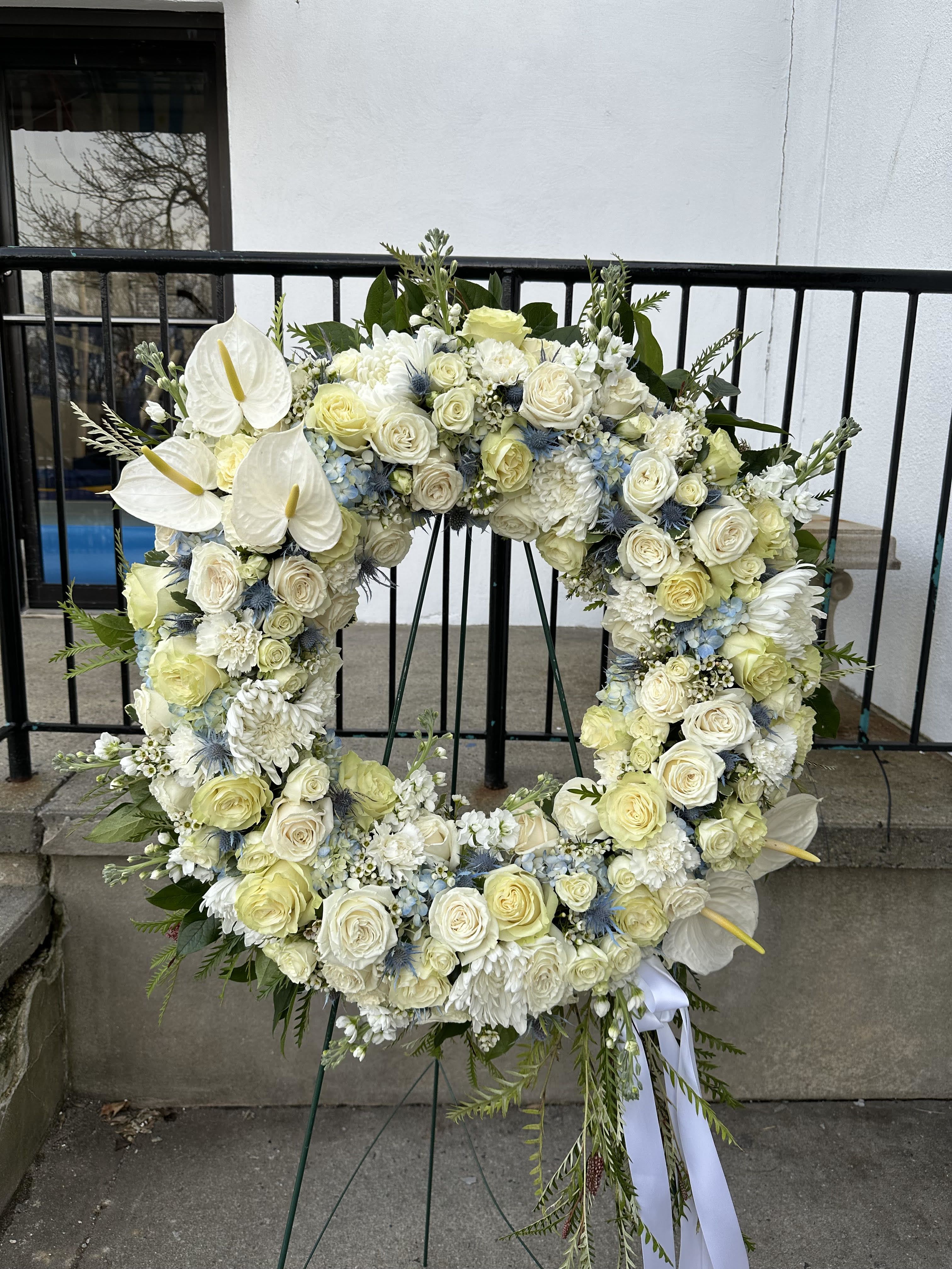 Designer Choice Sympathy Wreath - A graceful, hand-crafted wreath designed with a serene selection of white flowers accented by a gentle touch of blue. Thoughtfully arranged to honor a loved one's memory with elegance and peace. Available in Standard, Deluxe, and Premium sizes.