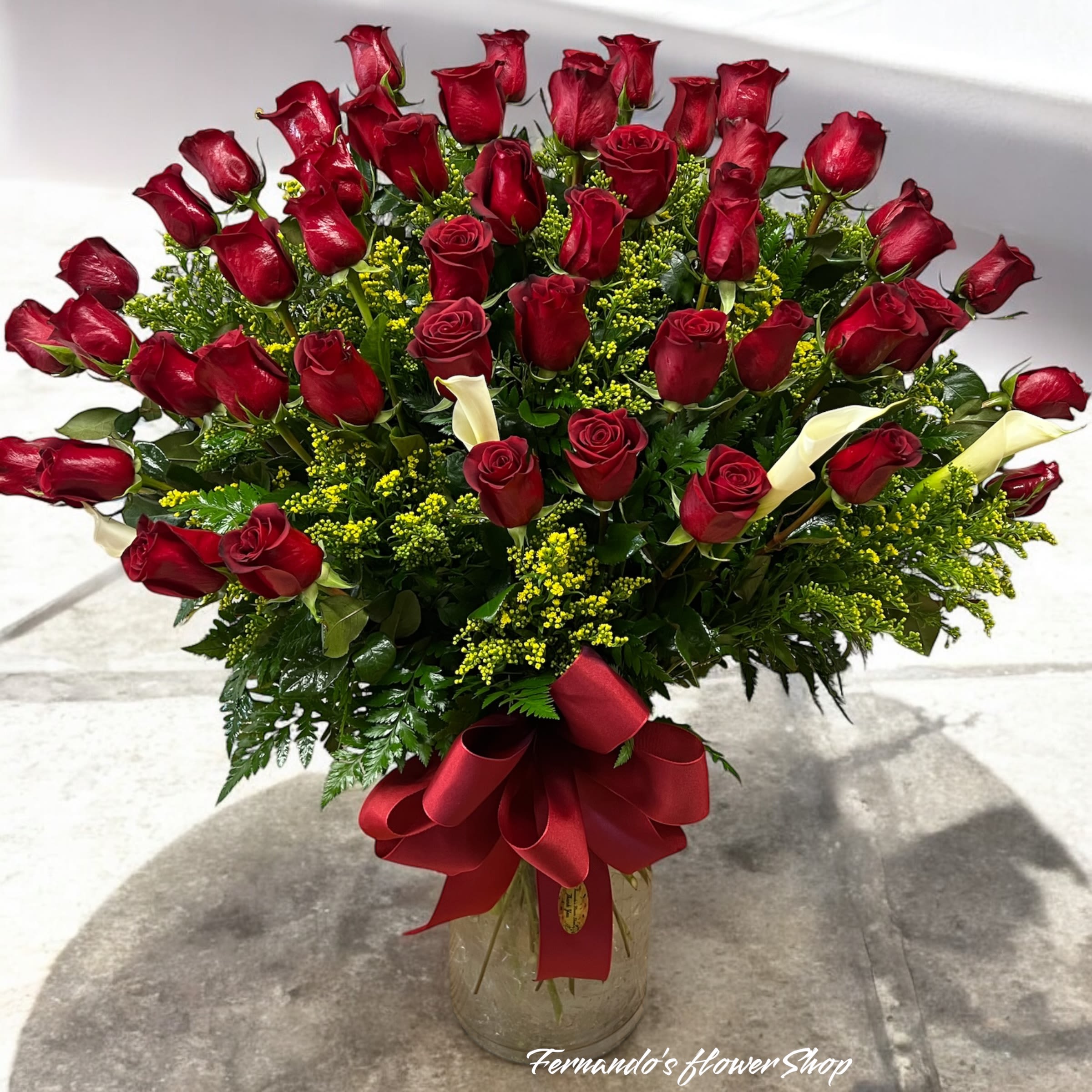 Timeless Bouquet - This image shows a beautifully arranged bouquet featuring a large number of red roses, accented with yellow filler flowers and a few white calla lilies. The bouquet is complemented by lush green foliage and is placed in a glass vase. The arrangement is adorned with a decorative red ribbon tied at the base, adding elegance to the display. The design appears to come from a florist, as indicated by the visible text “Fernando’s flowerShop” at the bottom of the image. 50 Roses….