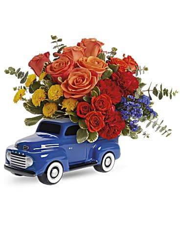 Ford Pick  - This image showcases a colorful floral arrangement placed in a decorative ceramic container shaped like a vintage blue pickup truck. The bouquet consists of roses in shades of orange and red, complemented by yellow and blue accent flowers, along with green foliage. The combination of warm and cool tones creates a striking contrast, making this arrangement a charming and nostalgic gift or centerpiece.
